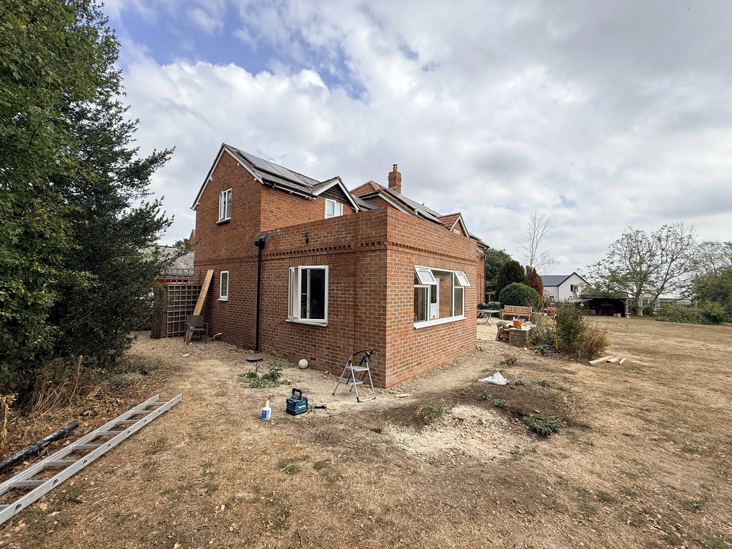 A house under construction with the back exterior brick wall partly built, exposing some interior work. Construction tools and materials are scattered on the dirt ground. Nearby trees and neighboring houses are also visible.