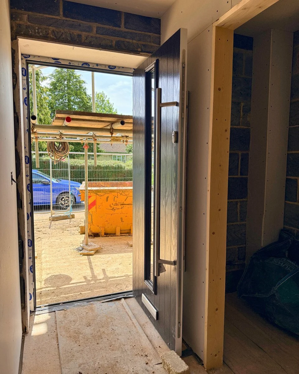View through an open door leading outside to a construction site with a blue car, a tree, and a house in the background. Inside, there are unfinished walls and construction materials.