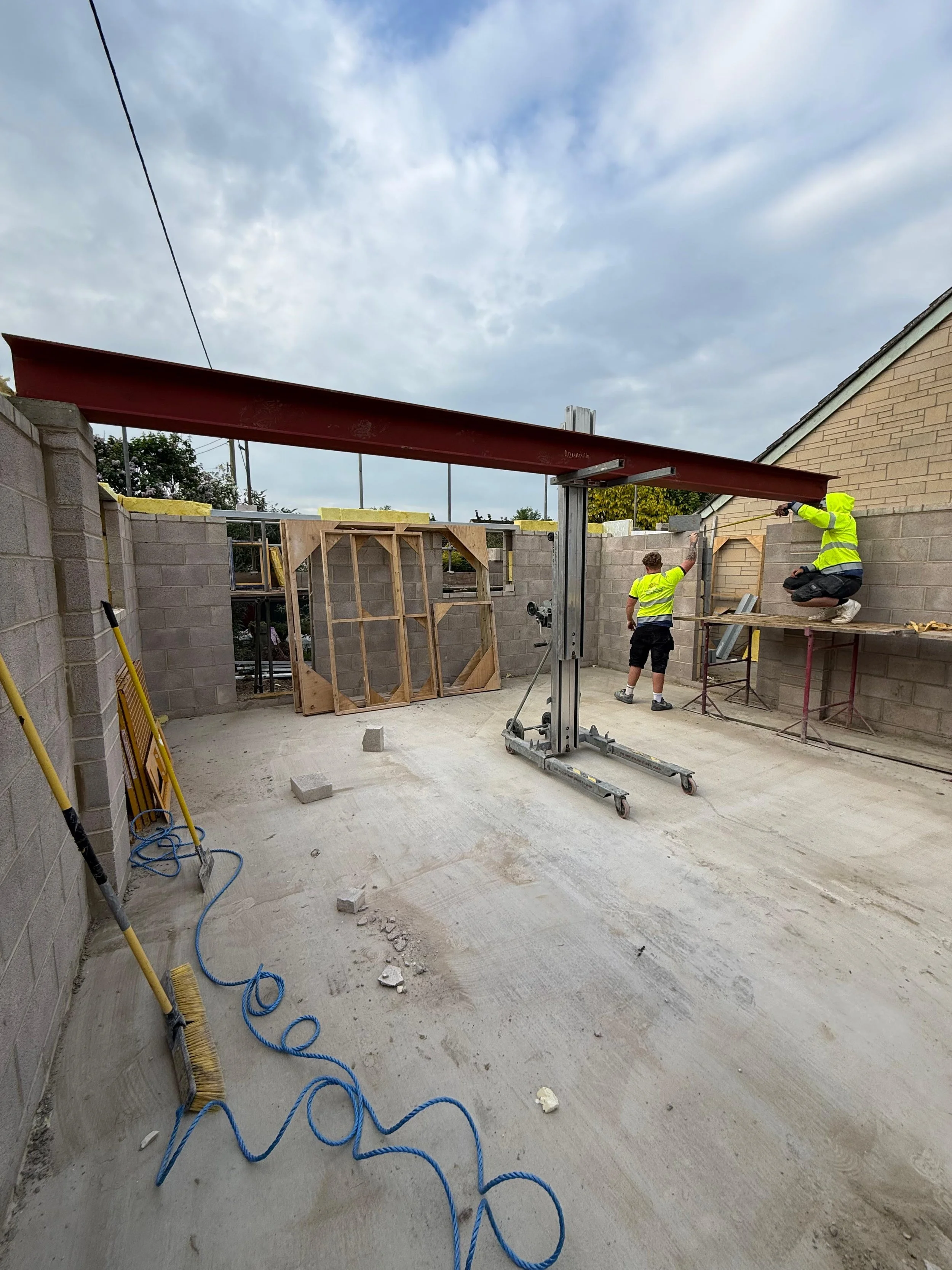 Construction workers in high visibility clothing working inside a building under construction, with steel beams and brick walls, and construction tools on the floor.