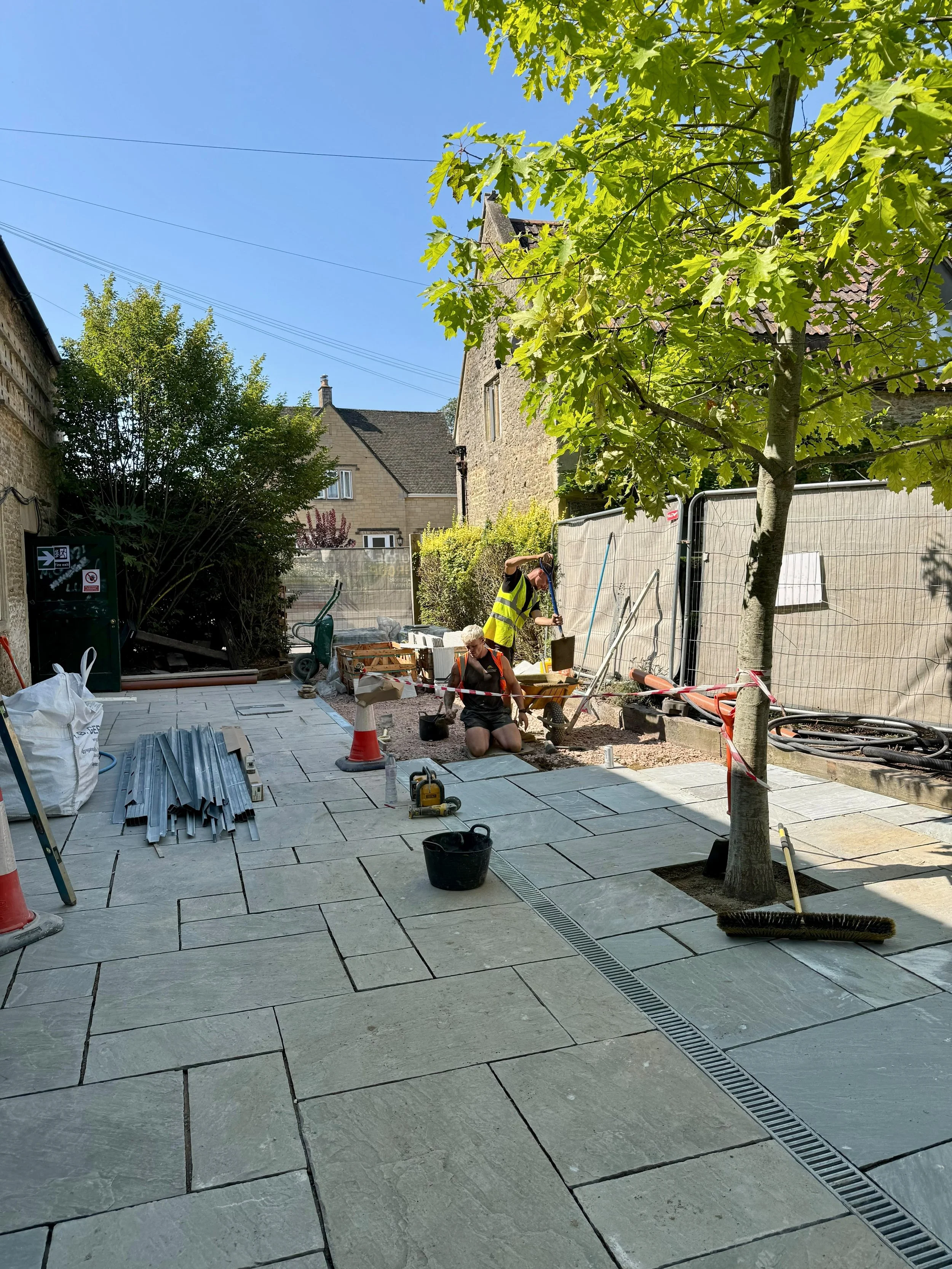 Workers installing paving stones on a sidewalk in a residential area, with construction materials and equipment around, a tree providing shade, and neighboring houses visible in the background.