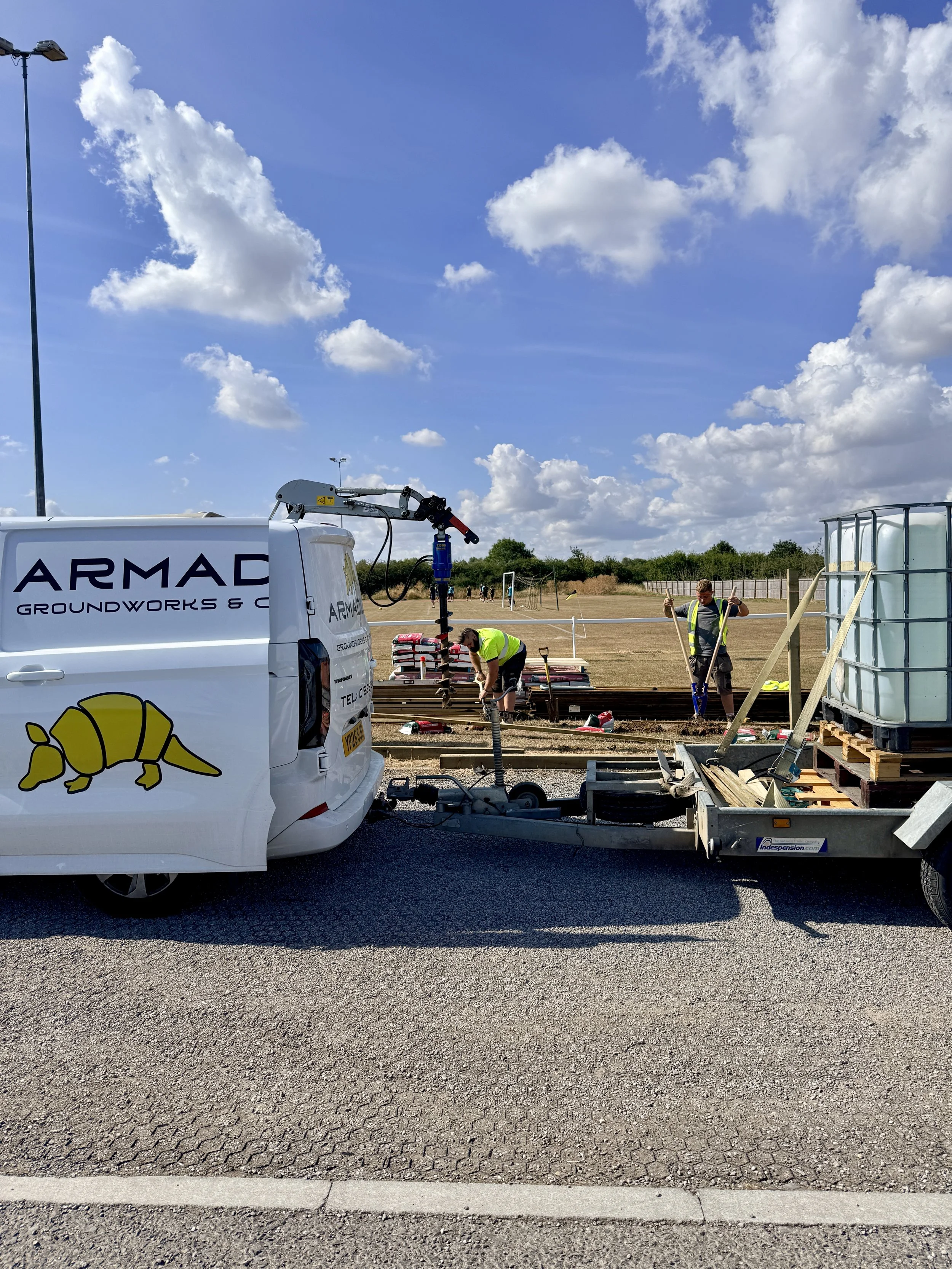 A construction site with two workers installing railroad tracks, with a white van labeled 'ARMAC Groundworks &' and a trailer in the foreground, and a grassy field with trees and a blue sky with clouds in the background.