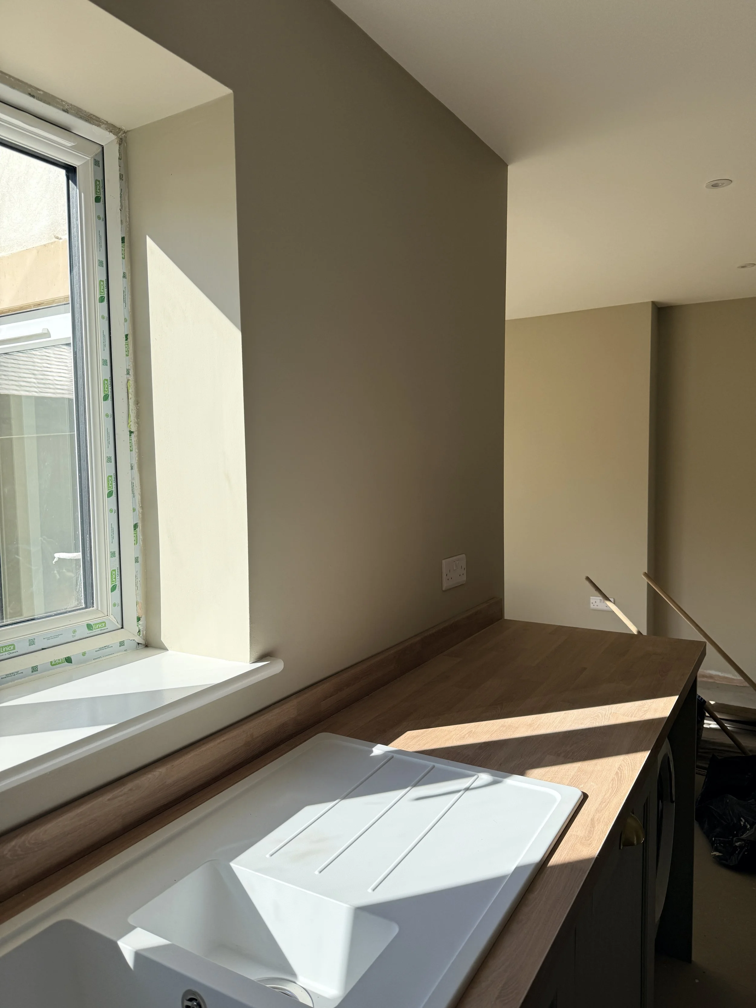 Corner of a kitchen with a white sink, wooden countertop, a window with a windowsill, and a beige wall with electrical outlets.
