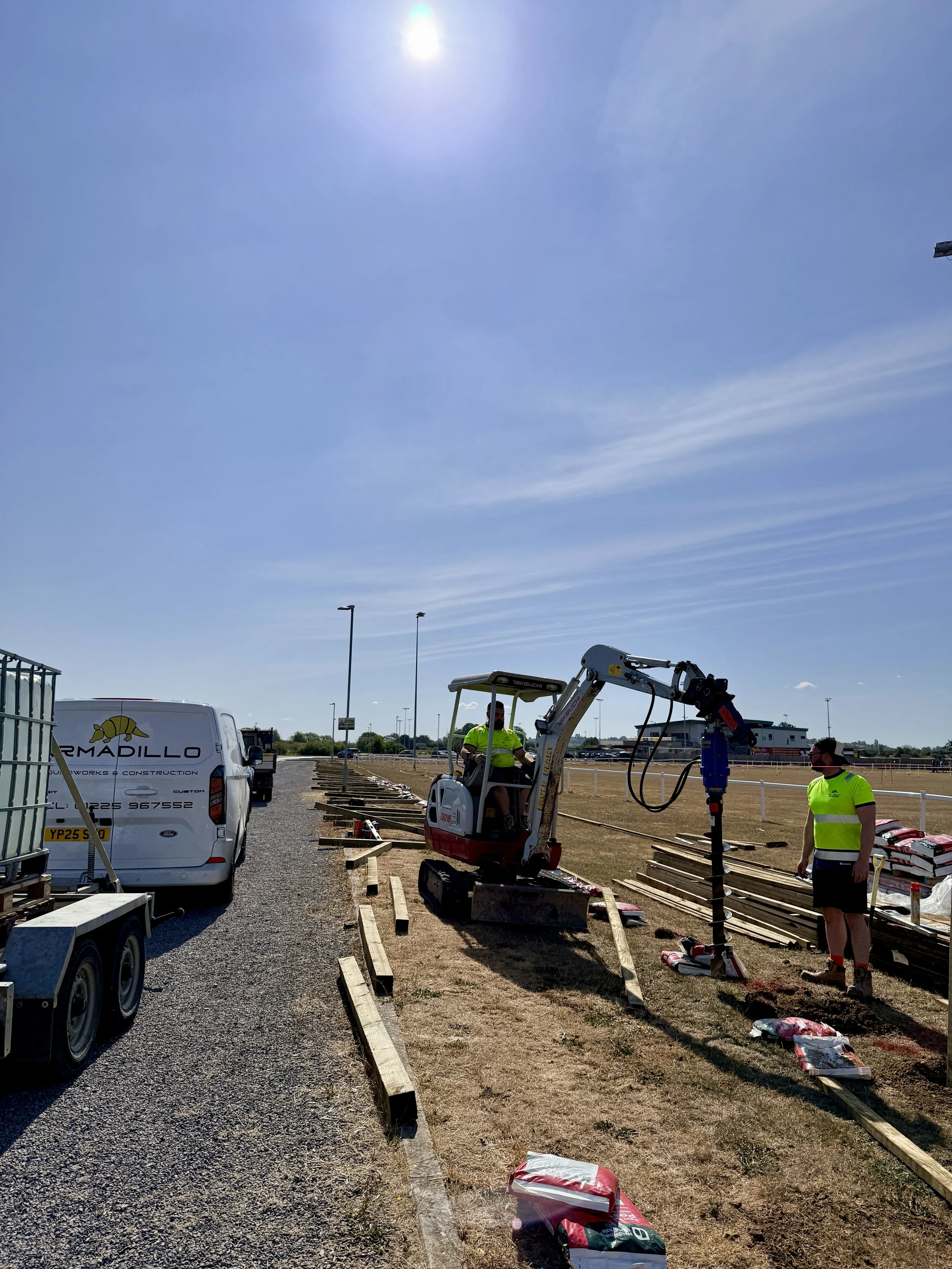 Construction workers installing a border or perimeter fence with wooden posts and supporting materials at an outdoor site on a sunny day.