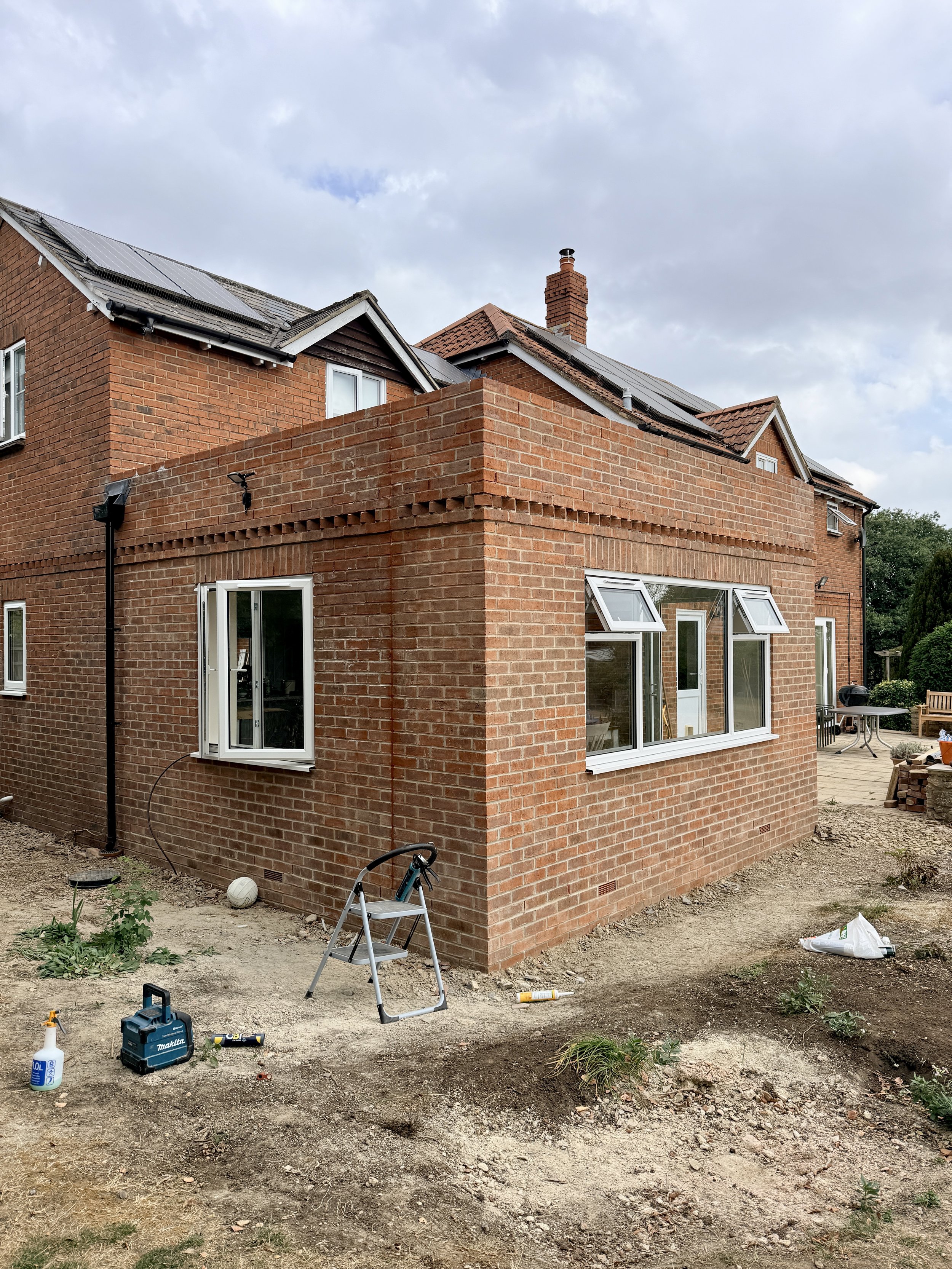 Backyard under construction with tools, plants, and building materials scattered on dirt ground, brick house with open windows, and cloudy sky