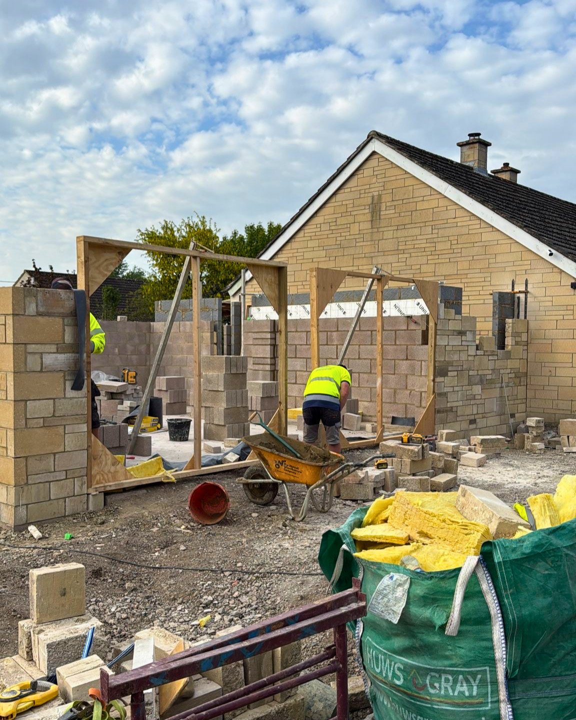 Construction workers building a brick wall at a house construction site with materials and tools scattered around.