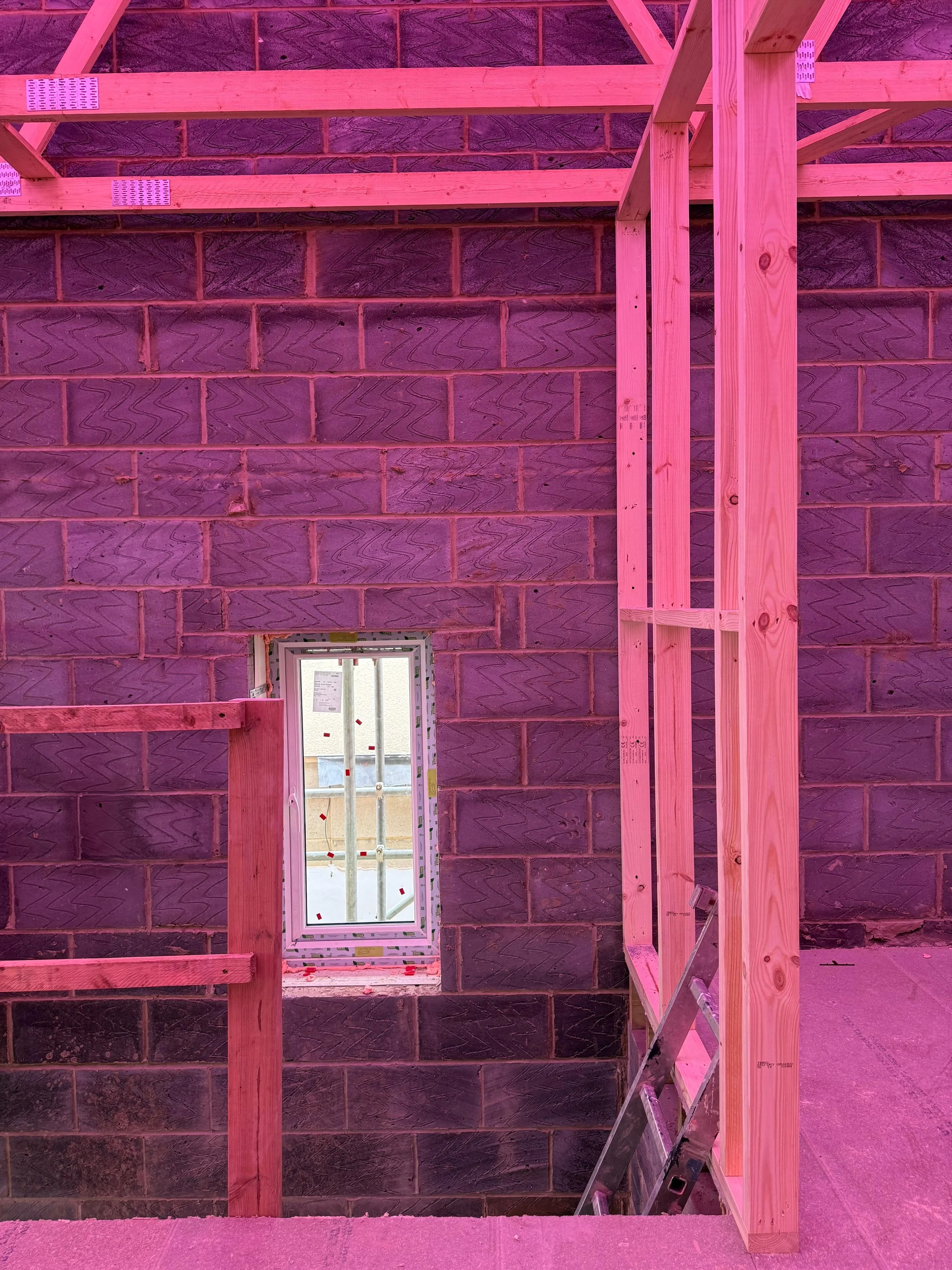 Interior view of a building under construction, with wooden framing and a small window in the brick wall. The lighting has a pink hue.