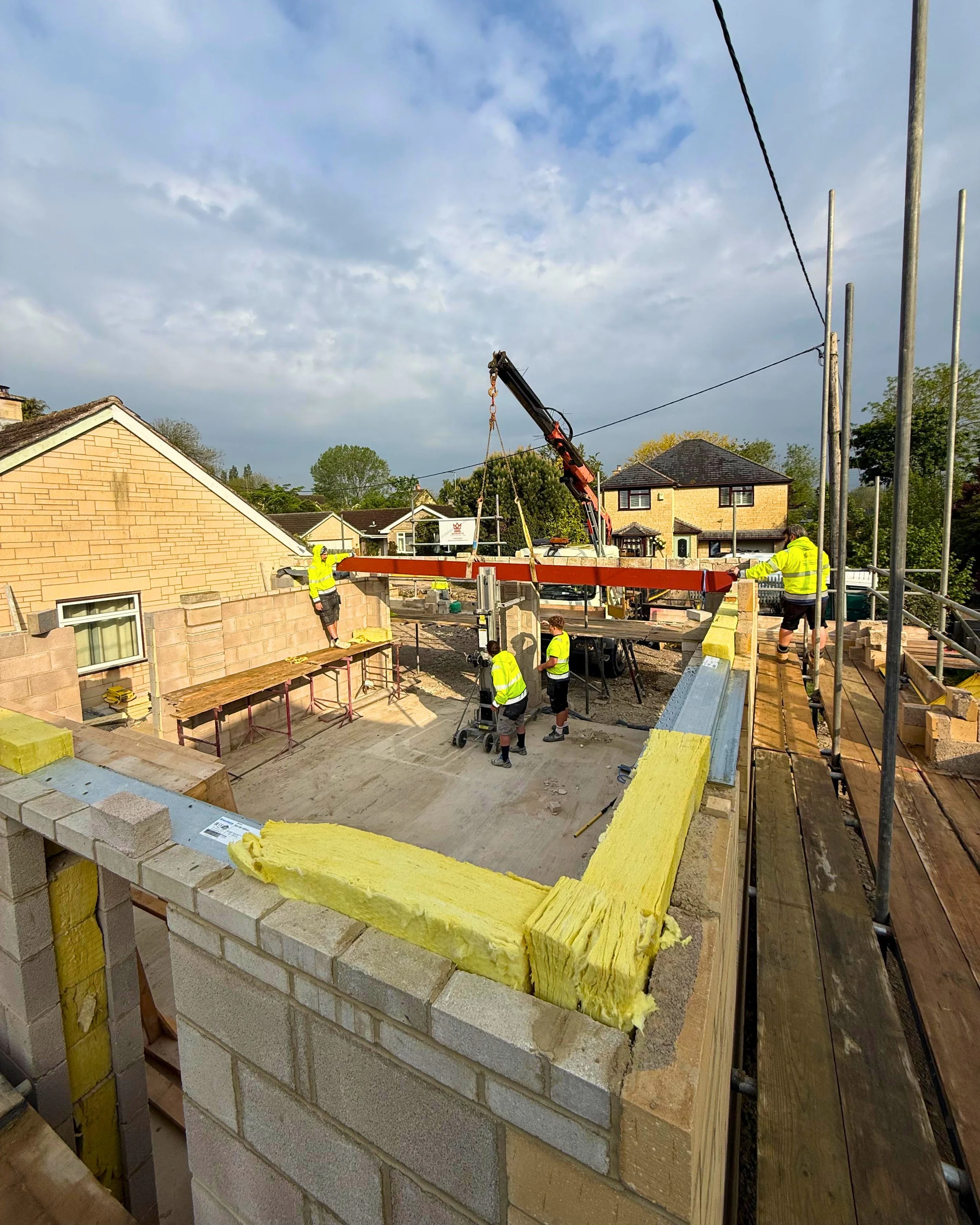 Construction workers building a brick wall with insulation material on top in a residential area. The workers are wearing safety vests and helmets. There is scaffolding and construction equipment around.
