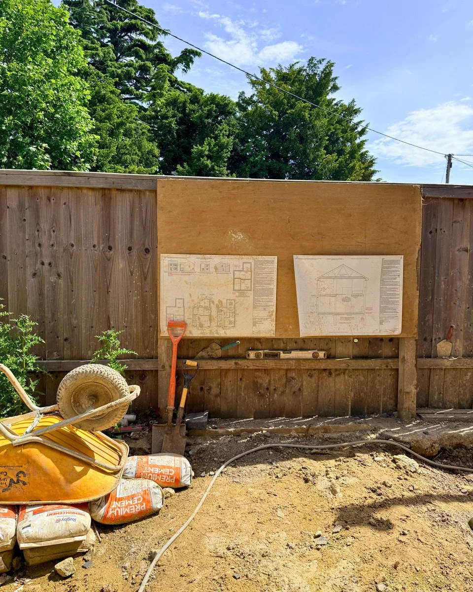 Backyard construction site with wooden fence, construction plans, wheelbarrow, bags of cement, and gardening tools. Green trees and blue sky in the background.