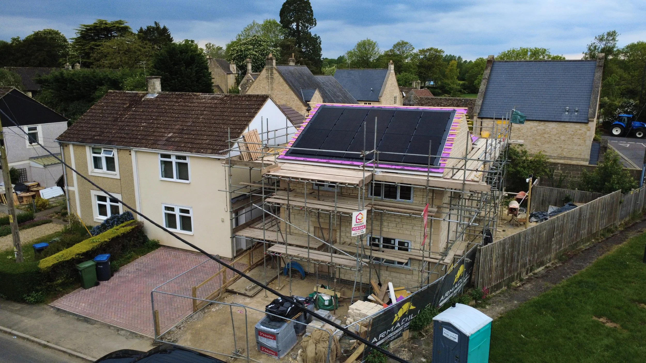A house under construction with scaffolding on its exterior, black solar panels on the roof, and surrounded by other houses and greenery.