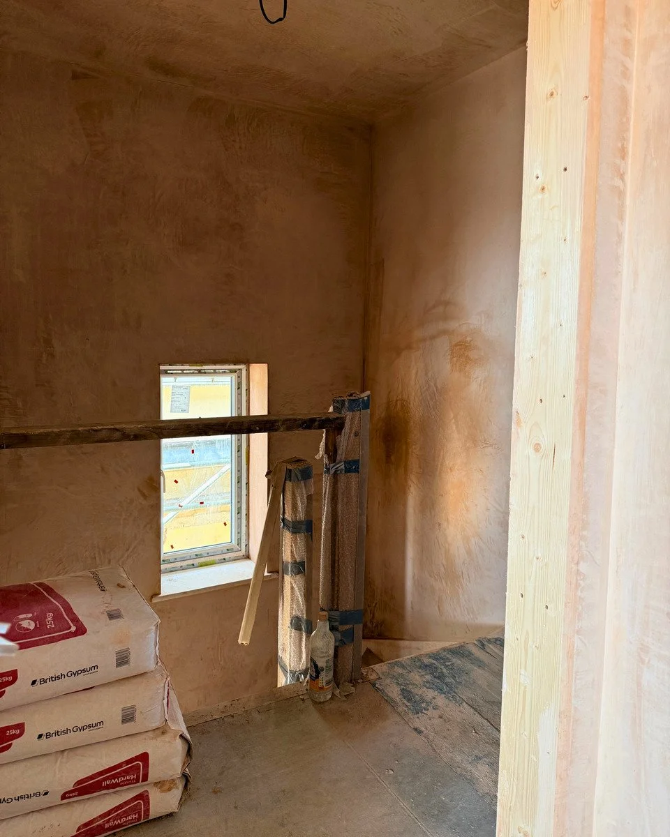 Interior of a room under construction with plastered walls, small window, and construction materials including bags of gypsum and tools.