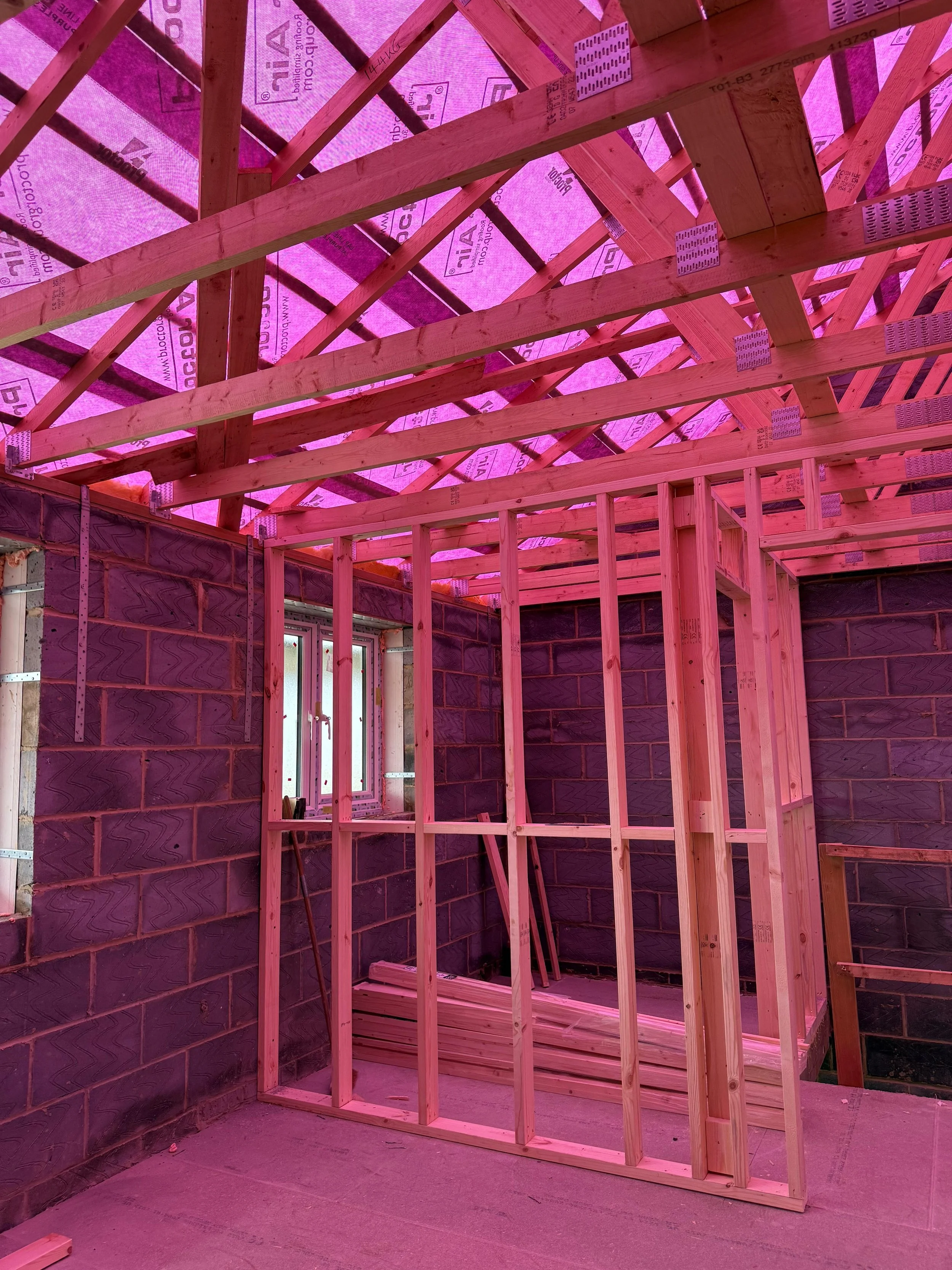Interior view of a building under construction, showing wooden framing, pink insulation on the ceiling, and a brick wall partially visible.