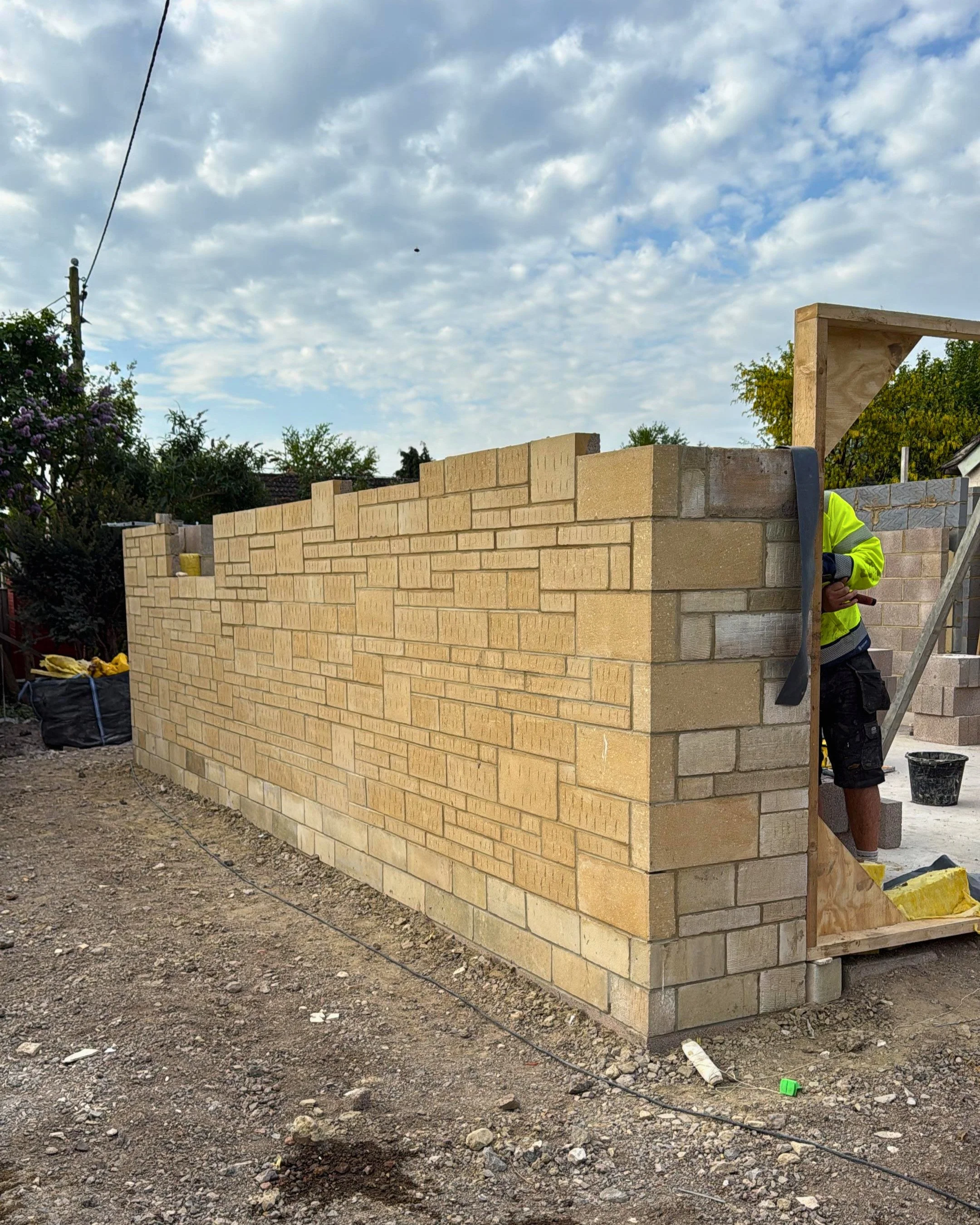 Construction site with beige brick wall partially built, worker in yellow safety jacket and black shorts standing beside it, cloudy sky overhead, tools and materials scattered on ground.