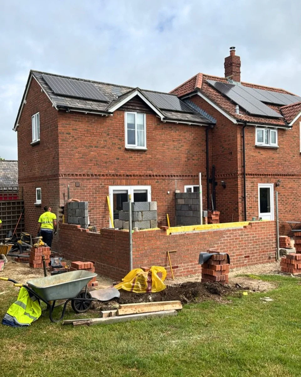 A two-story brick house under construction with workers present, building a small brick extension or wall at the back, outdoors on a cloudy day.