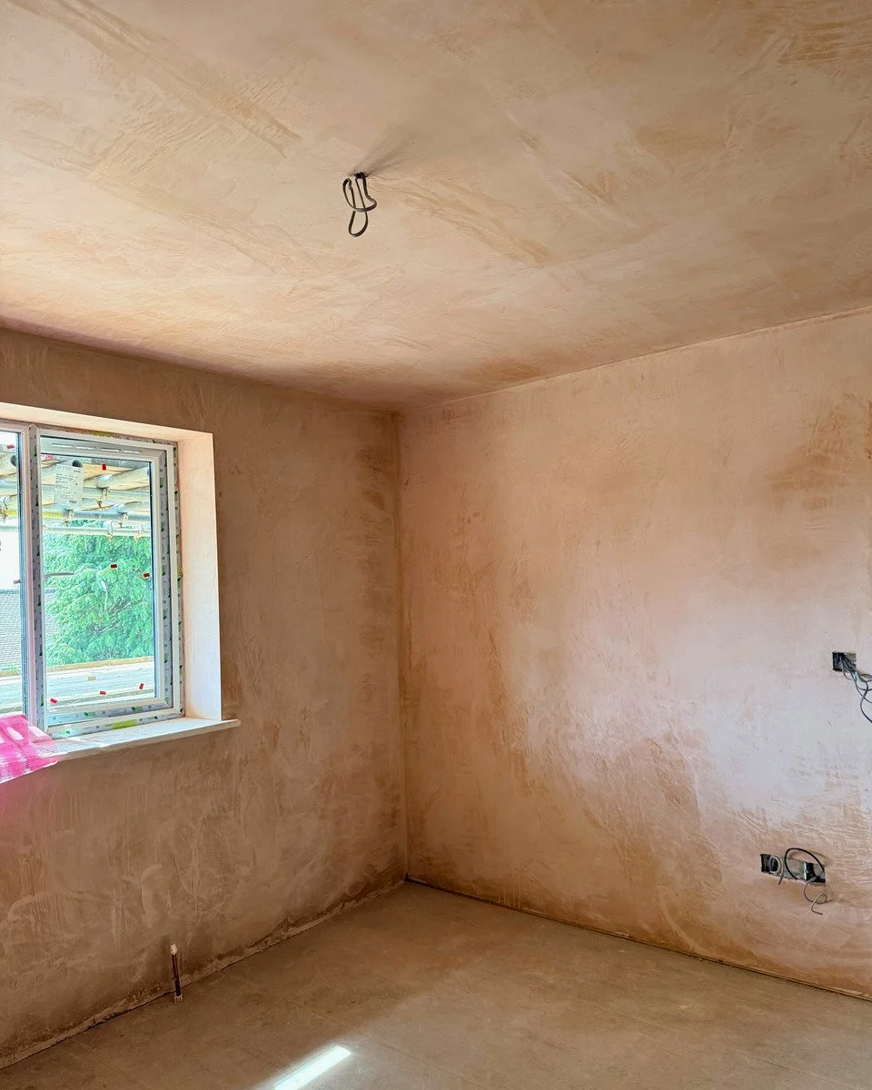 A room under construction with unfinished plaster walls and ceiling, a window with a view of green trees outside, and electrical wires and outlets not yet installed.
