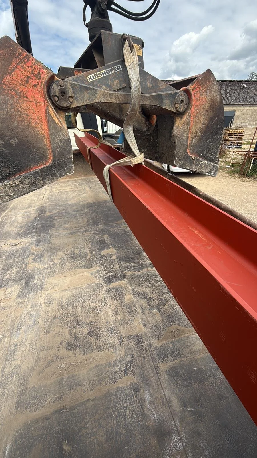 Close-up of a construction vehicle's arm attachment with a red metal beam secured to it, on a dusty worksite. Overcast sky with clouds and distant buildings in the background.