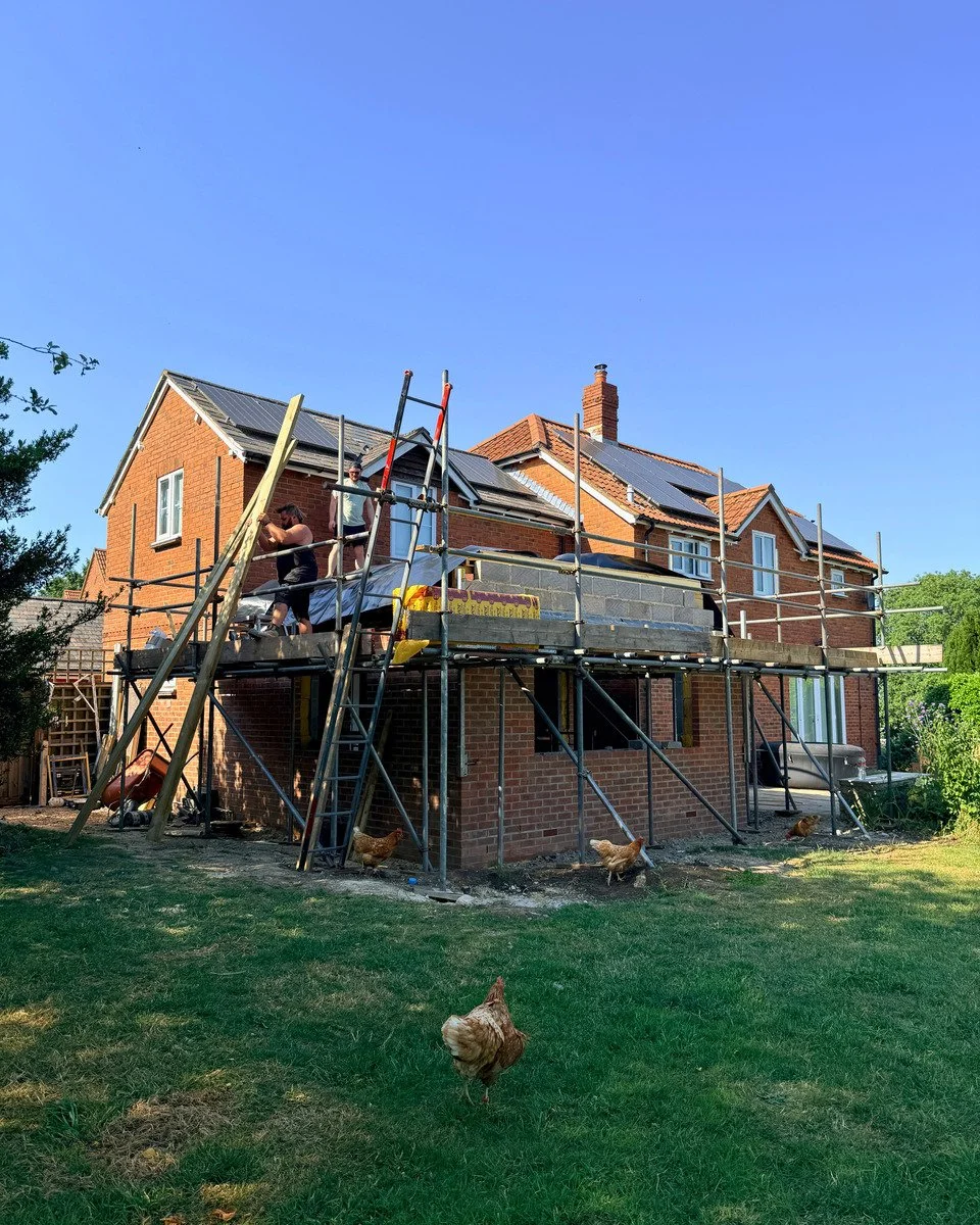 Construction workers building an extension on a brick house with scaffolding, while chickens are roaming in the yard.