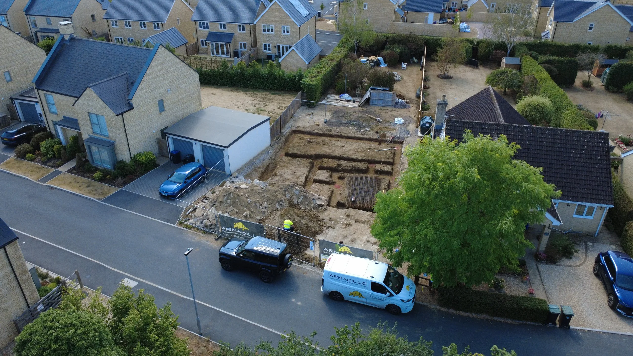 Aerial view of a house under construction with excavated trenches in the backyard and construction workers present, neighboring houses with parked cars visible.