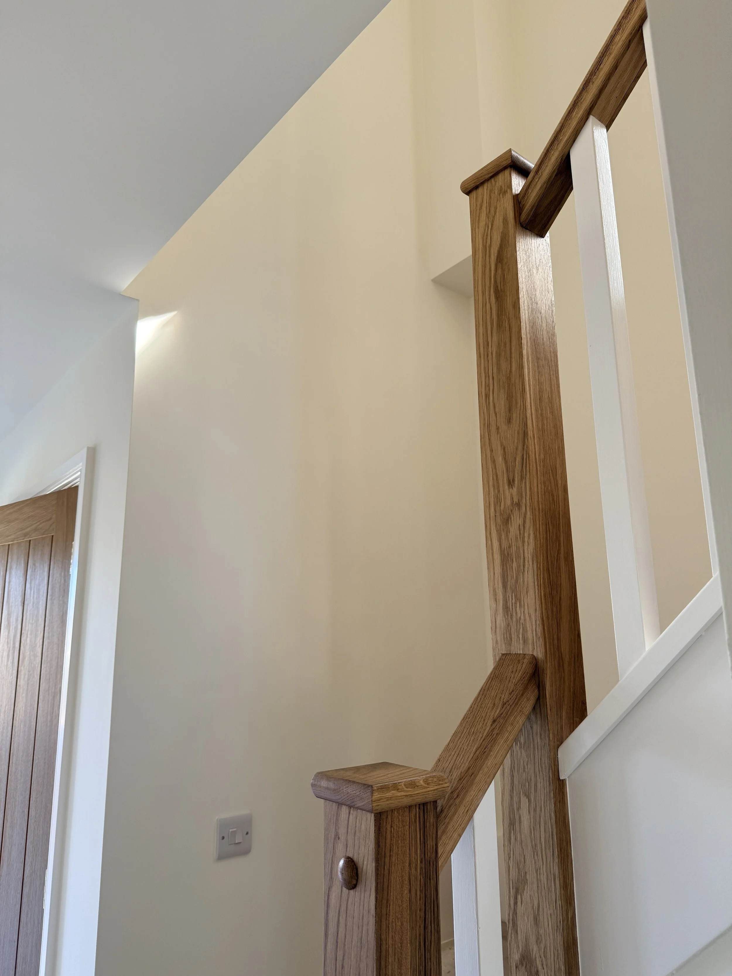 View of a wooden staircase handrail and newel post in a home, with cream-colored walls and a door with wooden paneling in the background.