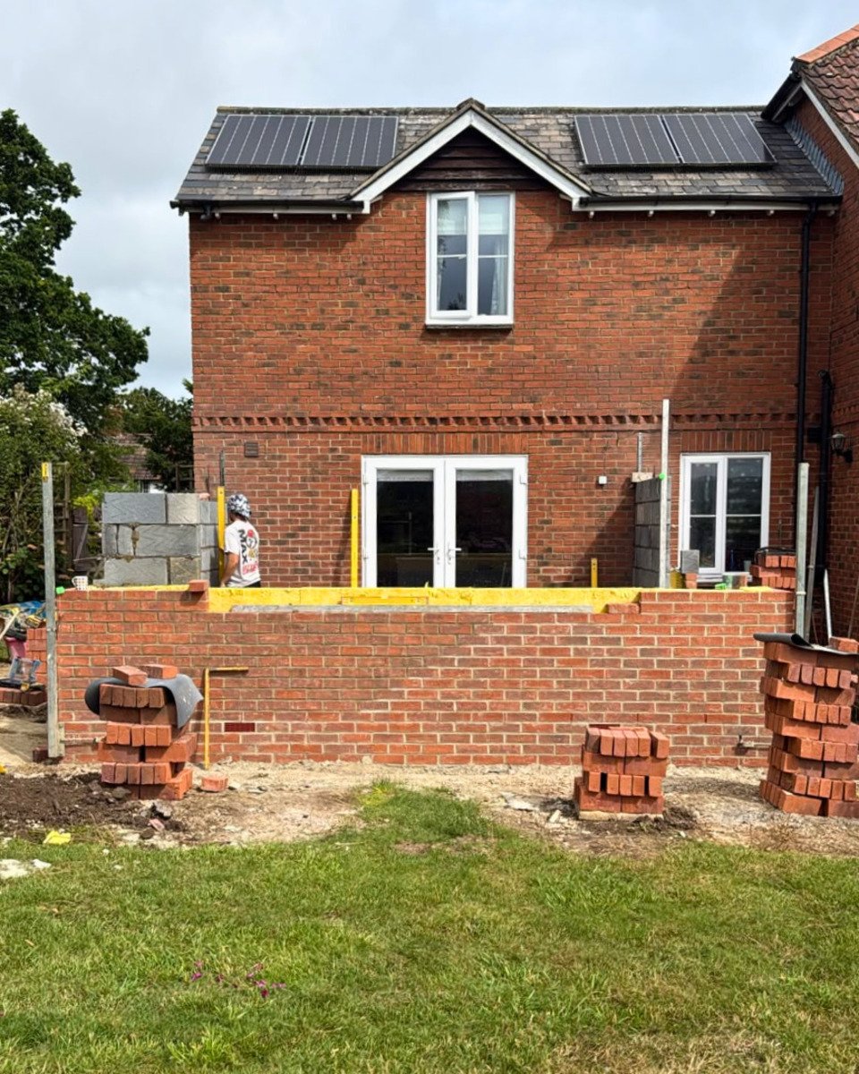 Brick house with solar panels on roof under construction, workers building a brick wall in backyard.