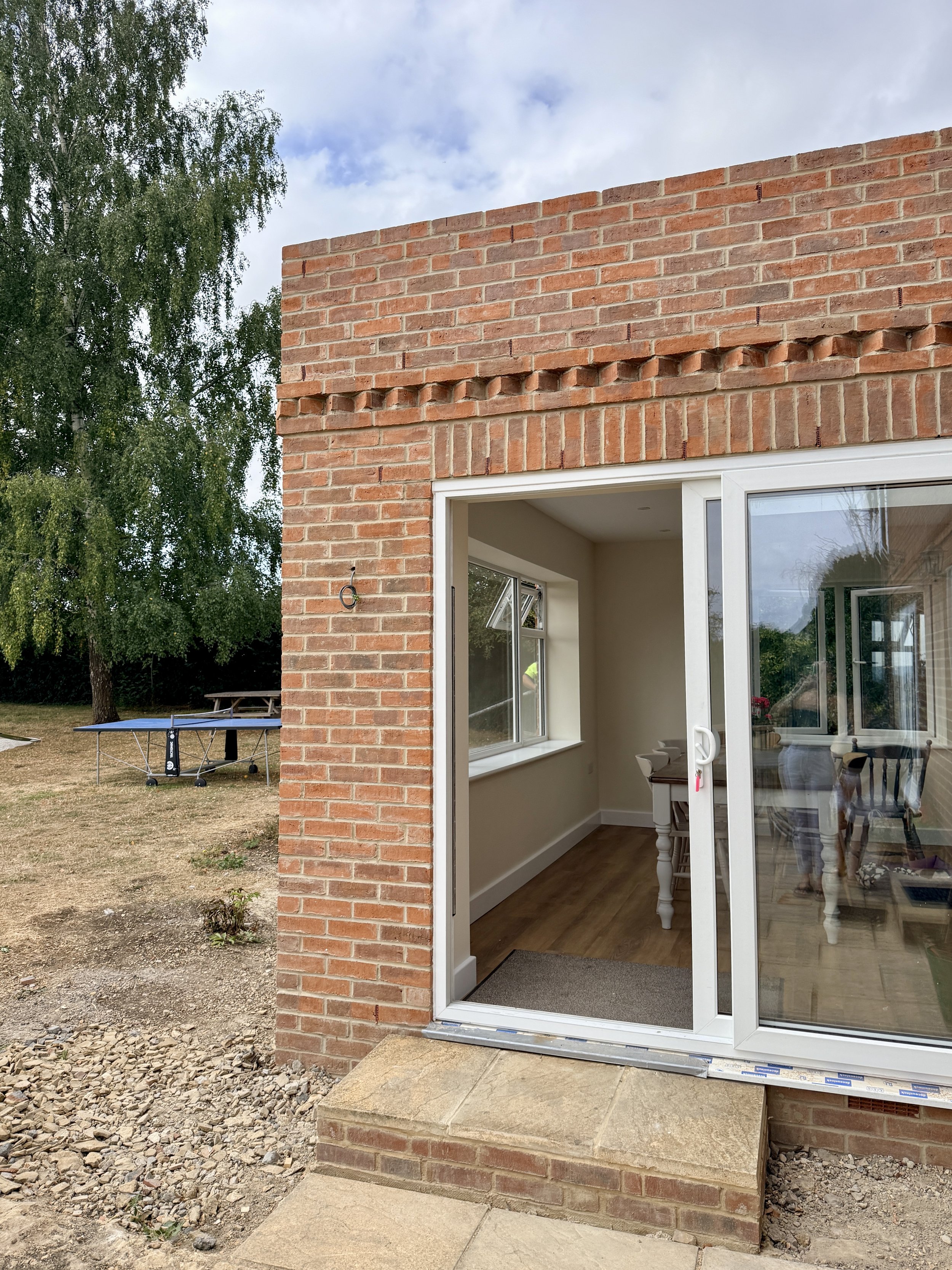 The image shows the exterior of a brick house with large glass sliding doors that open into a room with wooden flooring and chairs. There is a small set of steps leading up to the door. To the left, there is some bare soil and gravel, with a trampoli