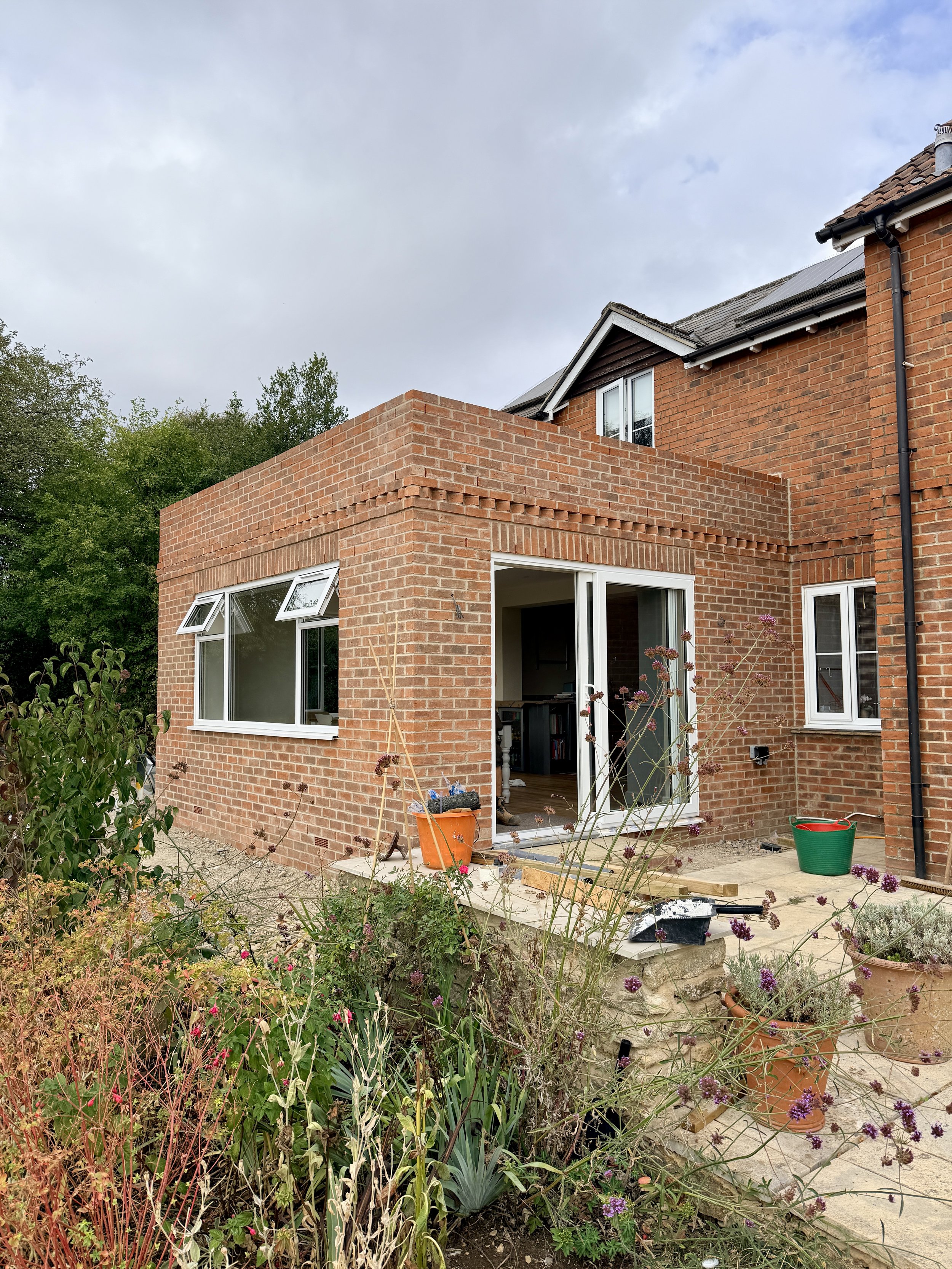 A two-story brick house under construction or renovation with an open sliding door leading to a room indoors. The house has a flat roof extension with large windows on the lower level. There are construction tools, buckets, and materials on the patio