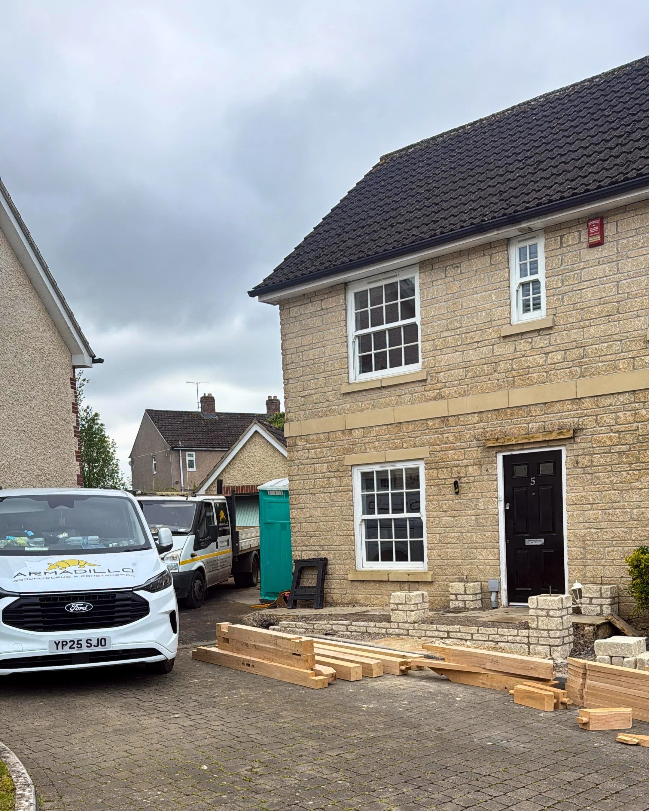 A residential house with yellow brick exterior and black front door, with cars parked in the driveway and construction materials, including wooden beams and bricks, in front of the house under a cloudy sky.