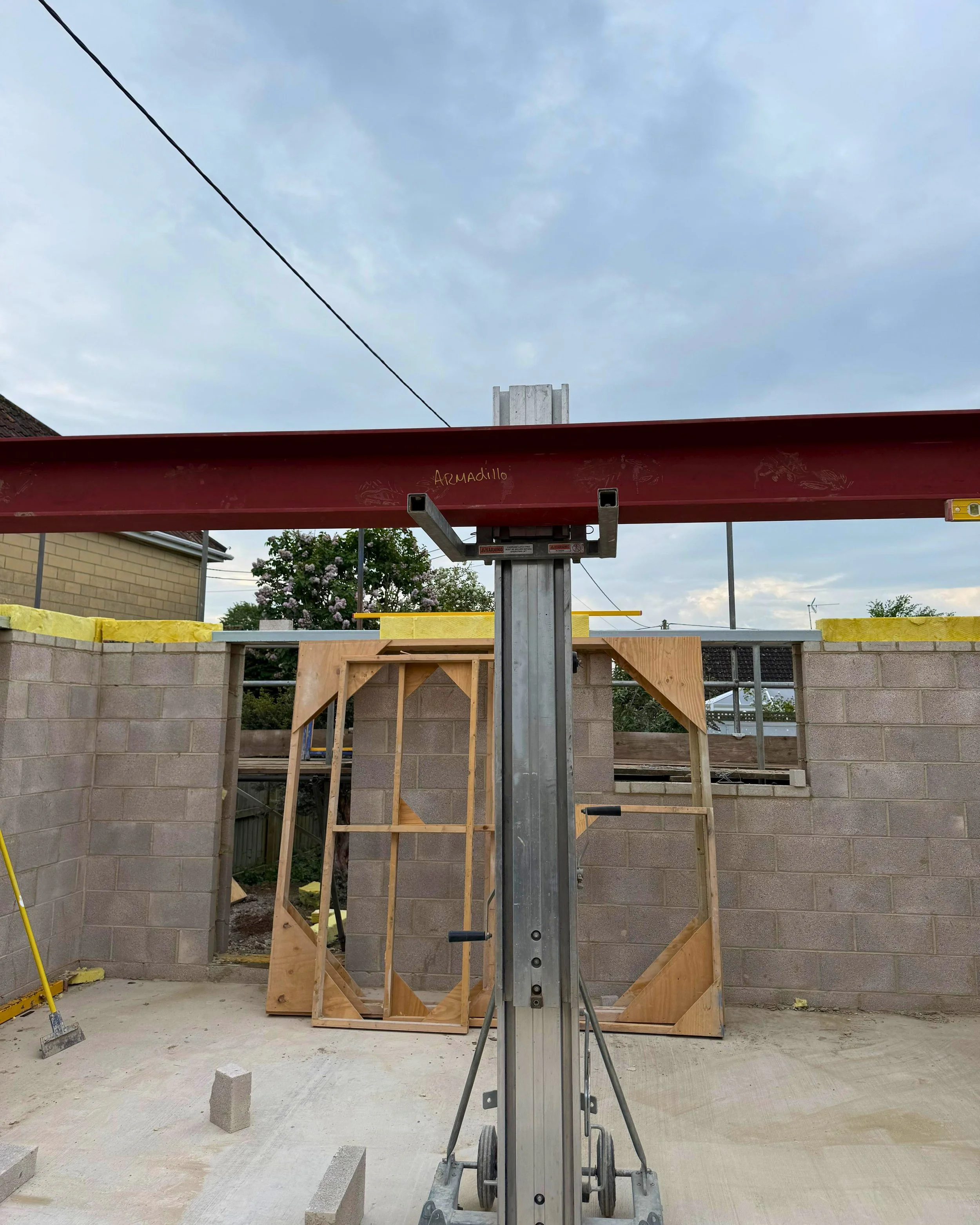 Construction site with framing for walls, a cement block wall, plywood frames, and a hydraulic lift tower in the center, under a cloudy sky.