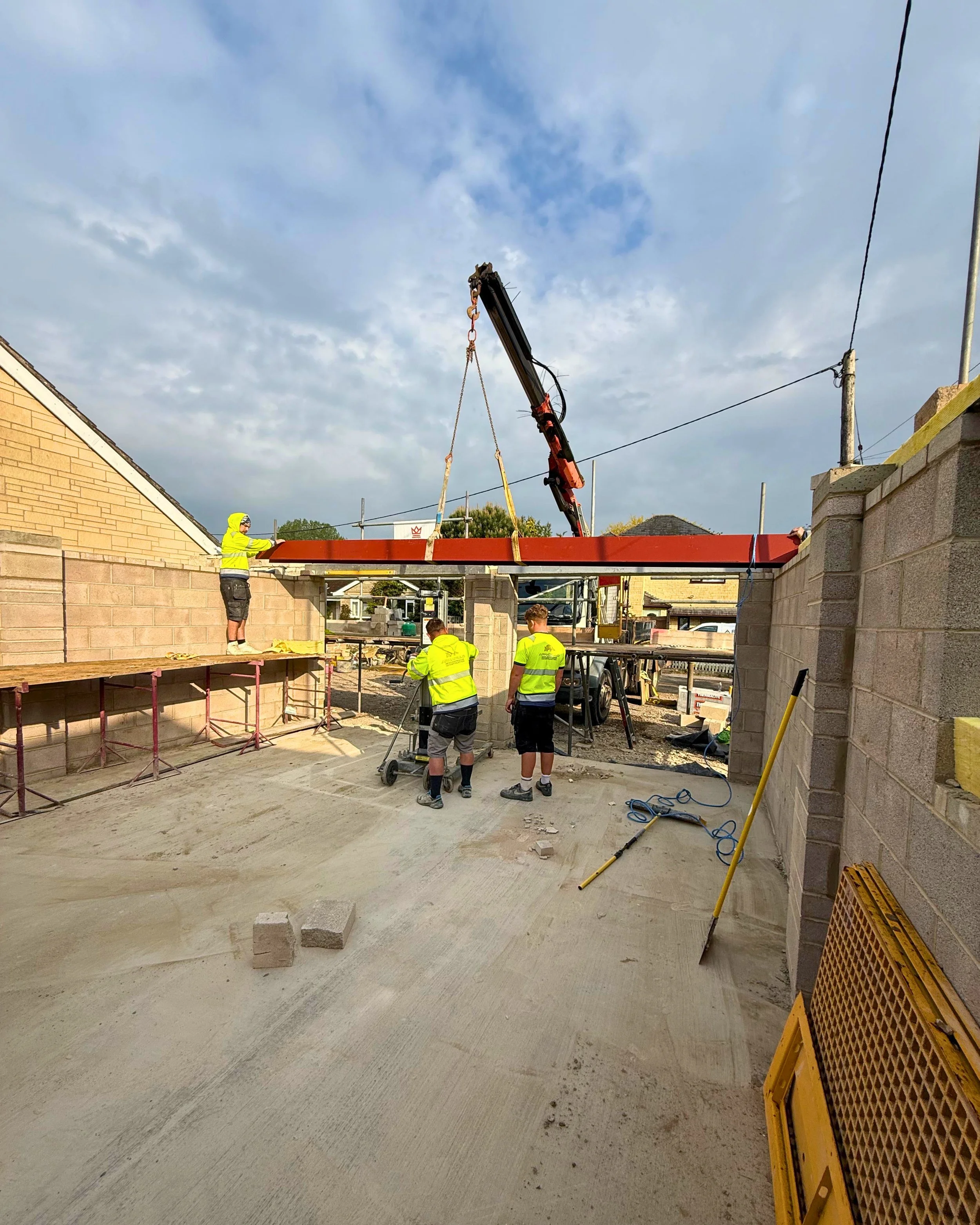 Construction workers in safety vests working on building a structure, with a crane lifting a beam and materials scattered on the ground.