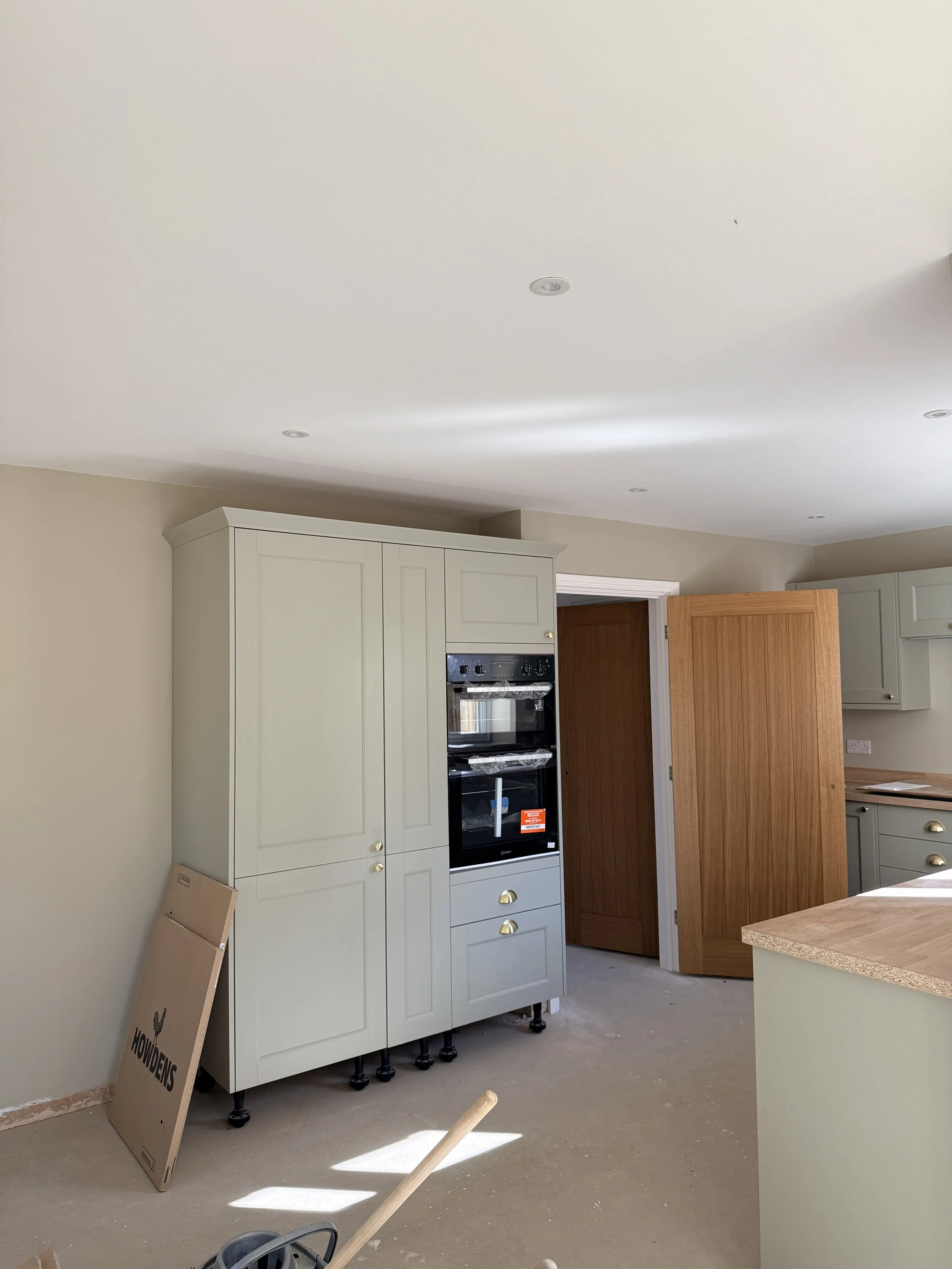 Kitchen room with beige walls, completed ceiling with recessed lights, pale green cabinetry, black double oven, wooden pantry door, cardboard box on floor, and a kitchen island with a beige top.