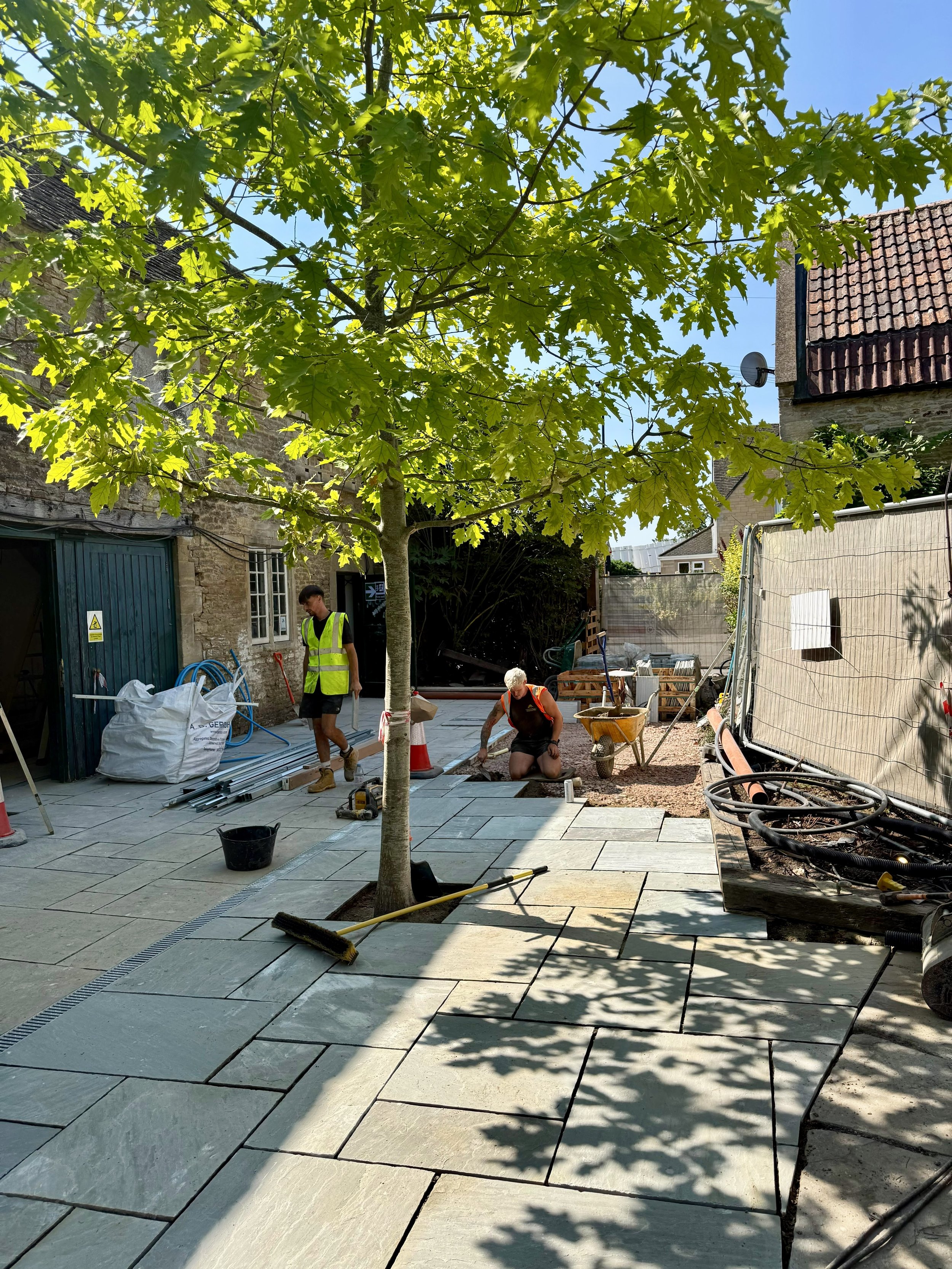Construction workers installing large stone or concrete tiles on a backyard patio, with a tree in the foreground and a small building with a tiled roof in the background.