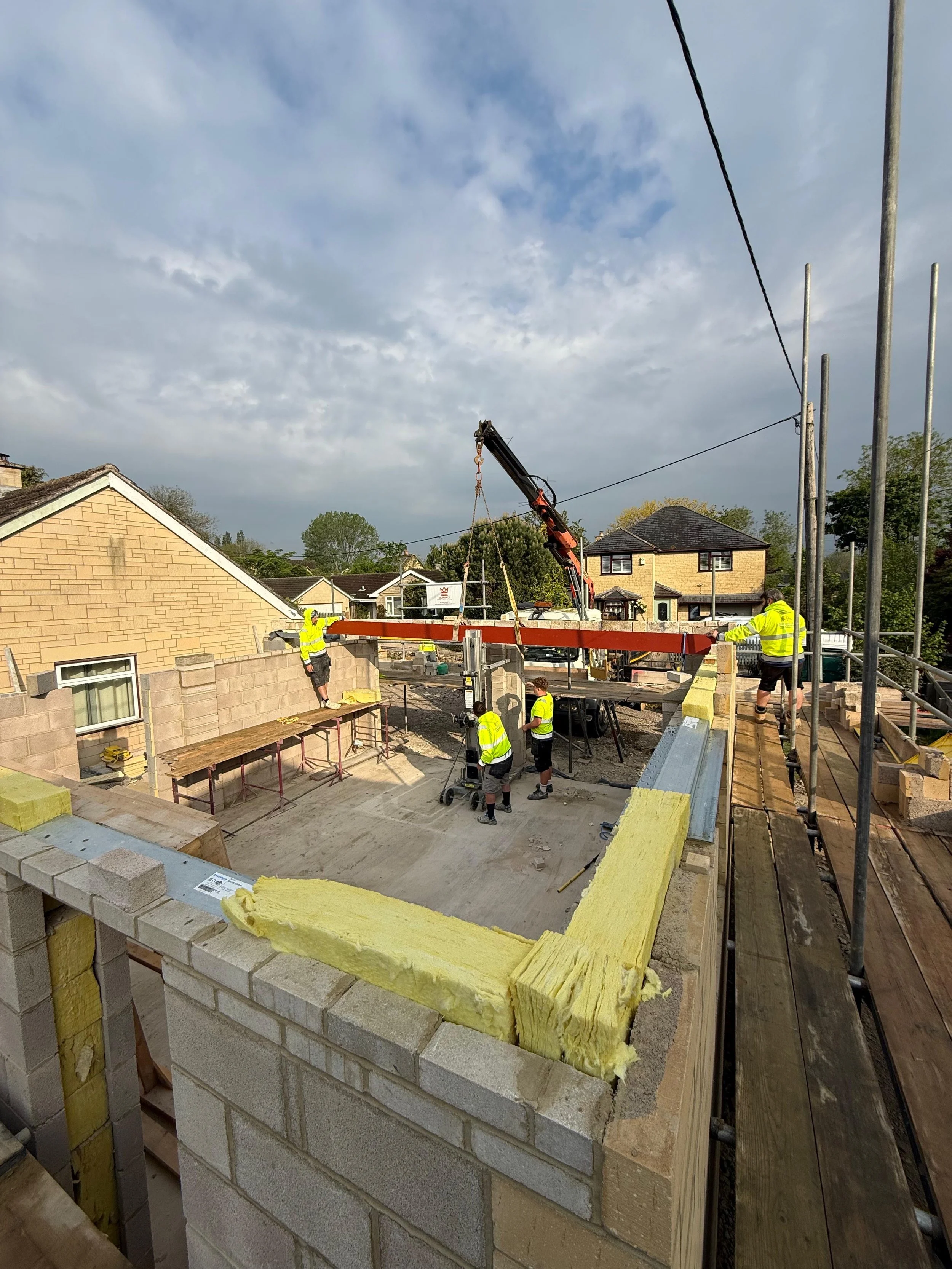 Construction workers building a house, with a crane lifting construction materials, surrounded by scaffolding and partially built brick walls.