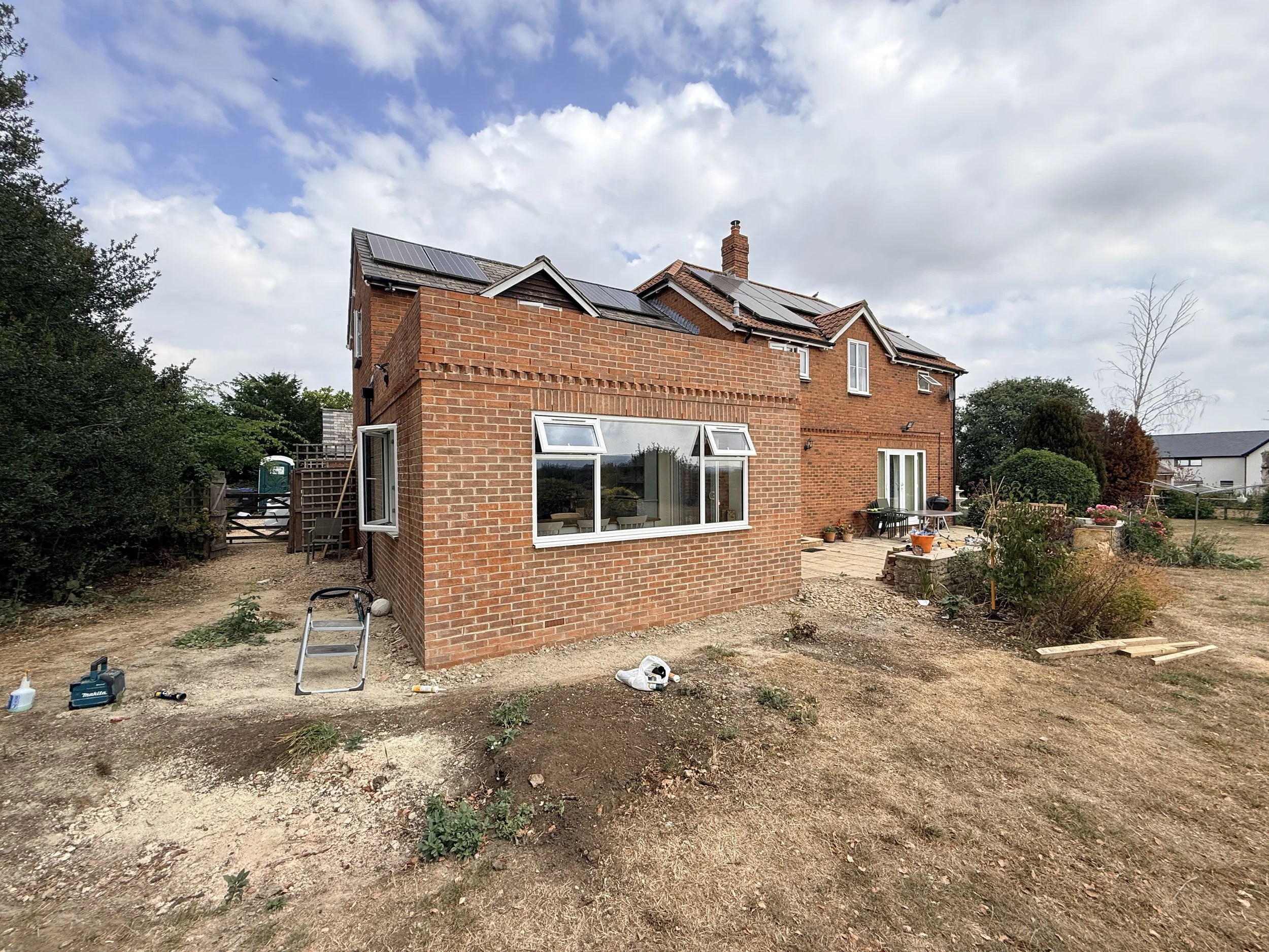 A two-story brick house under renovation with solar panels on the roof. The backyard has a dirt ground with construction tools and materials scattered around, including a step ladder, power tools, and a pile of wood. The house features large windows 