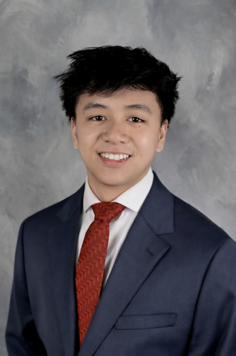 Young man in a brown suit, white shirt, and red tie standing outdoors with trees and a building in the background.