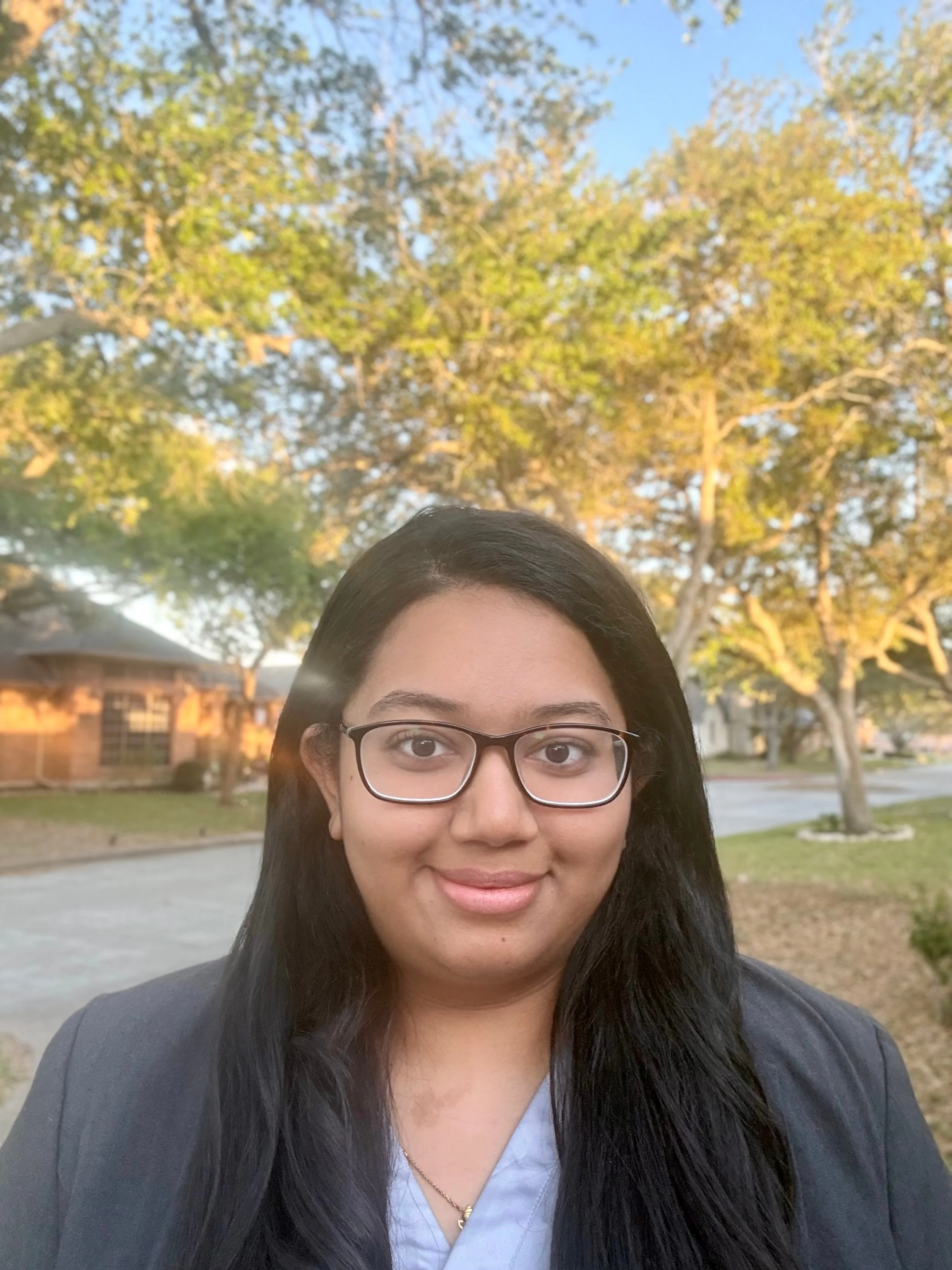 A young woman with glasses and long black hair taking a selfie outdoors, with trees and houses in the background during late afternoon or early evening.