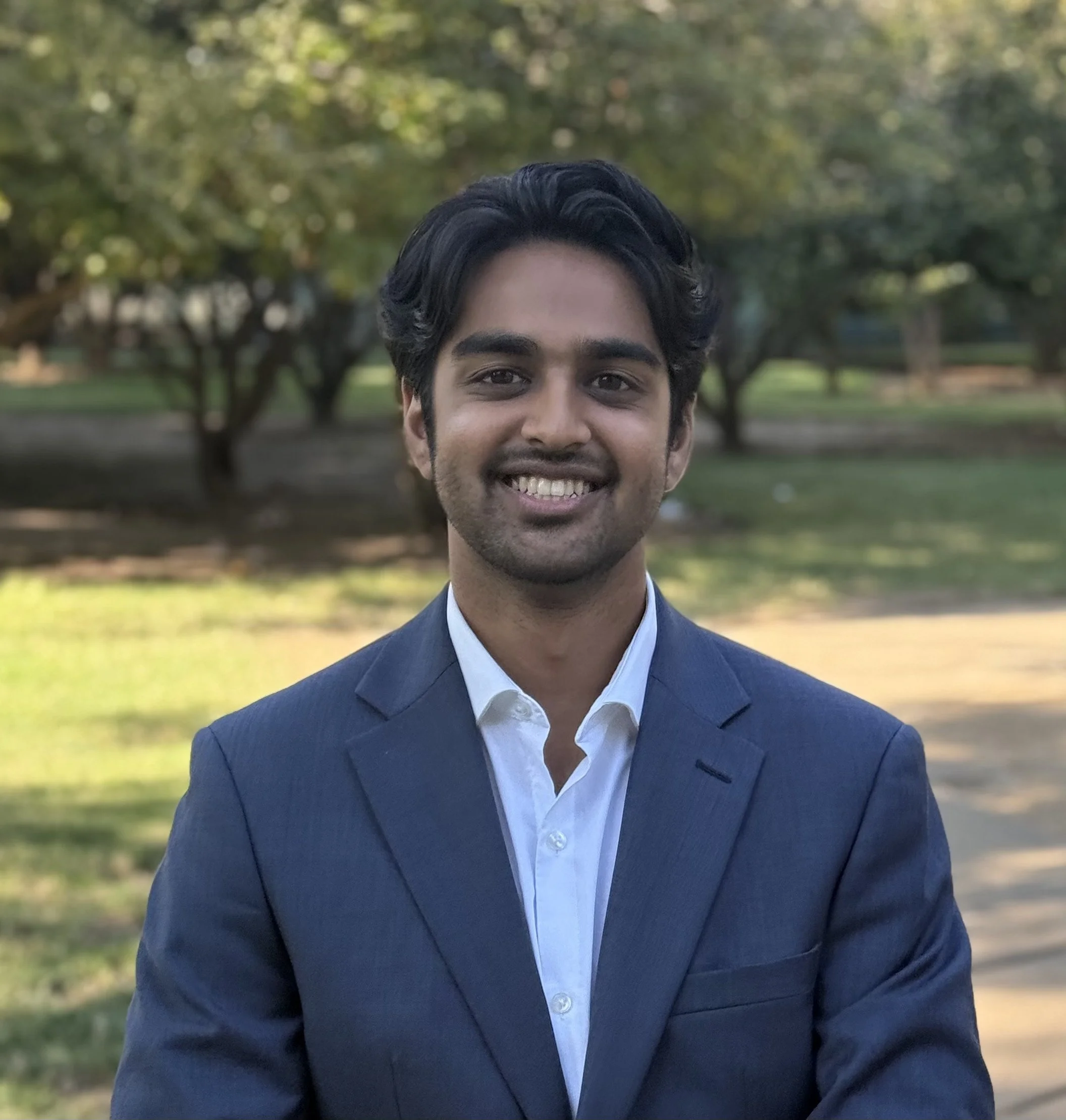 A young man in a blue suit and tie standing outdoors in front of a historic building with columns, smiling at the camera.