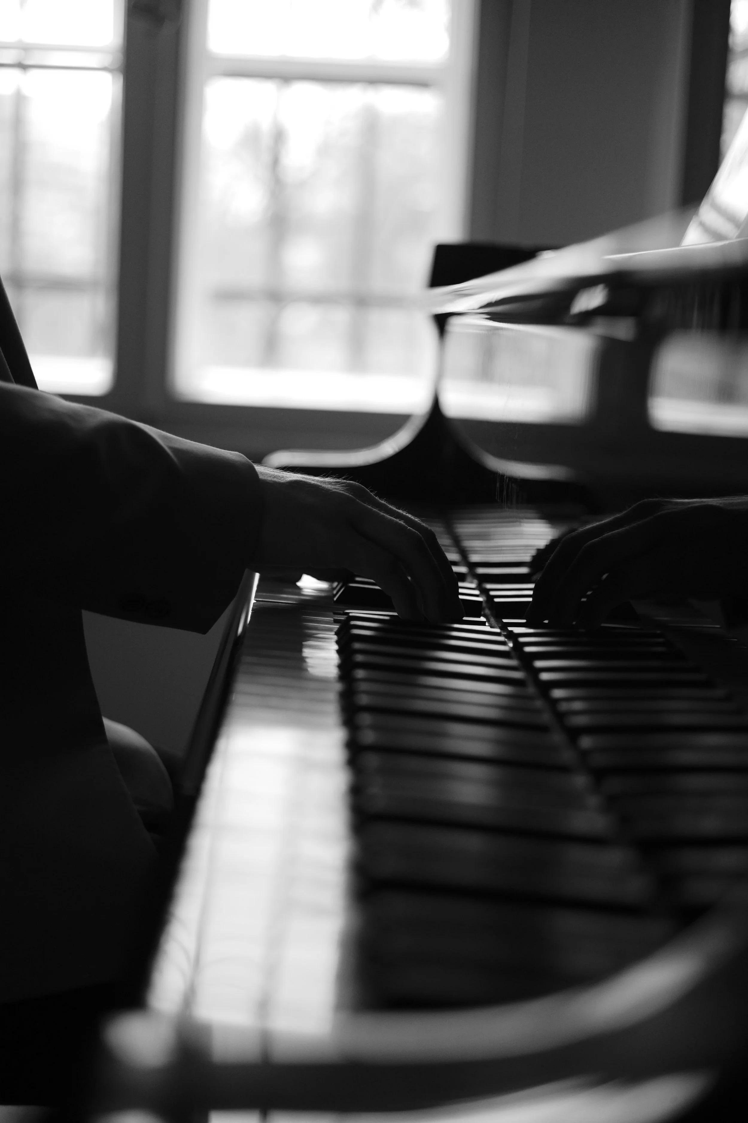 Black and white photo of a person playing a piano, with hands on the keys, near a window.