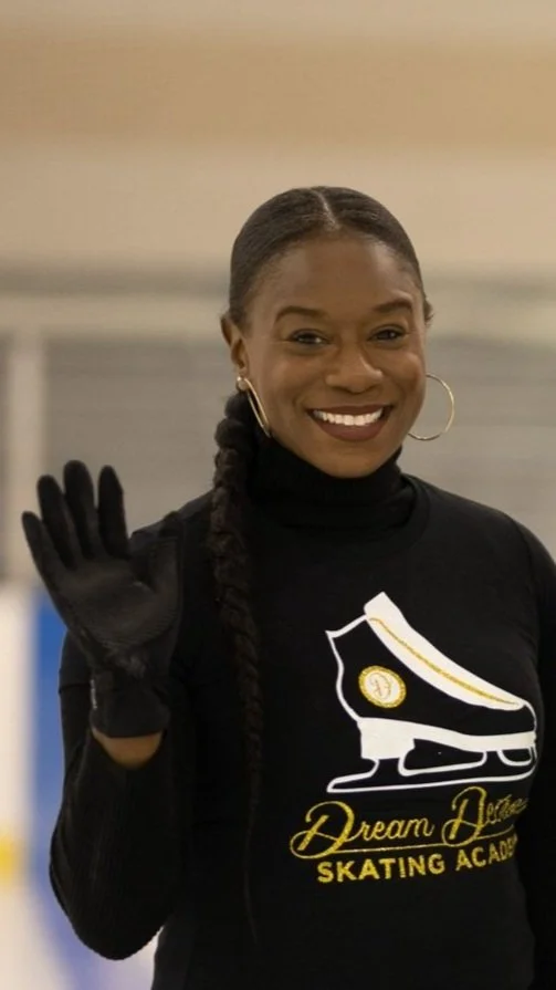 A smiling woman with dark hair in a braid, wearing a black long-sleeve shirt with a skating rink logo, gold hoop earrings, and black gloves, waving at the camera.