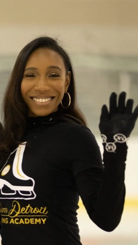 Woman smiling at an ice skating rink, wearing a black shirt, hoop earrings, and black gloves with bracelets.