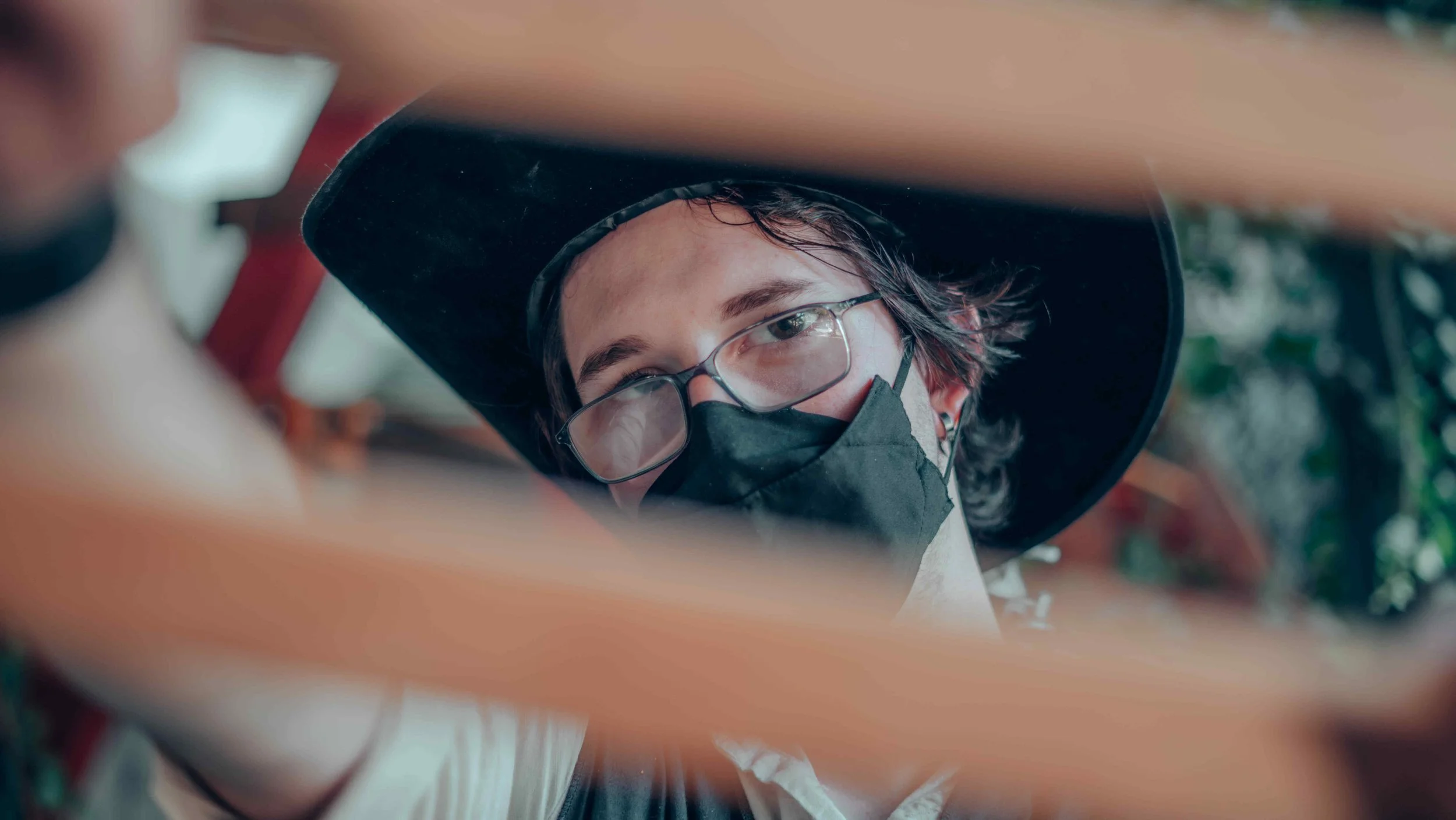 A person wearing glasses, a black face mask, and a black hat with a wide brim, looking downwards.