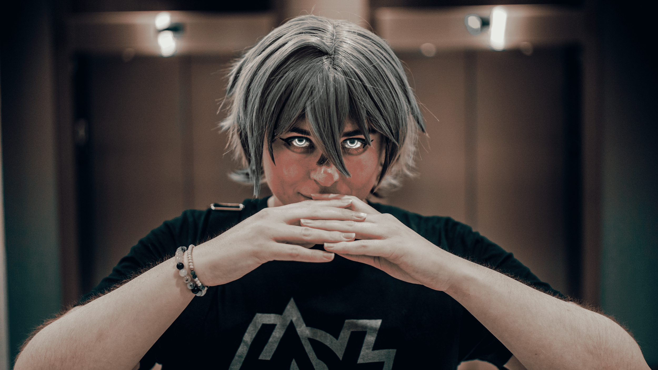 Person with short gray hair and dark eyeliner, resting chin on folded hands, wearing a black t-shirt with white text, in an indoor setting with blurred background.