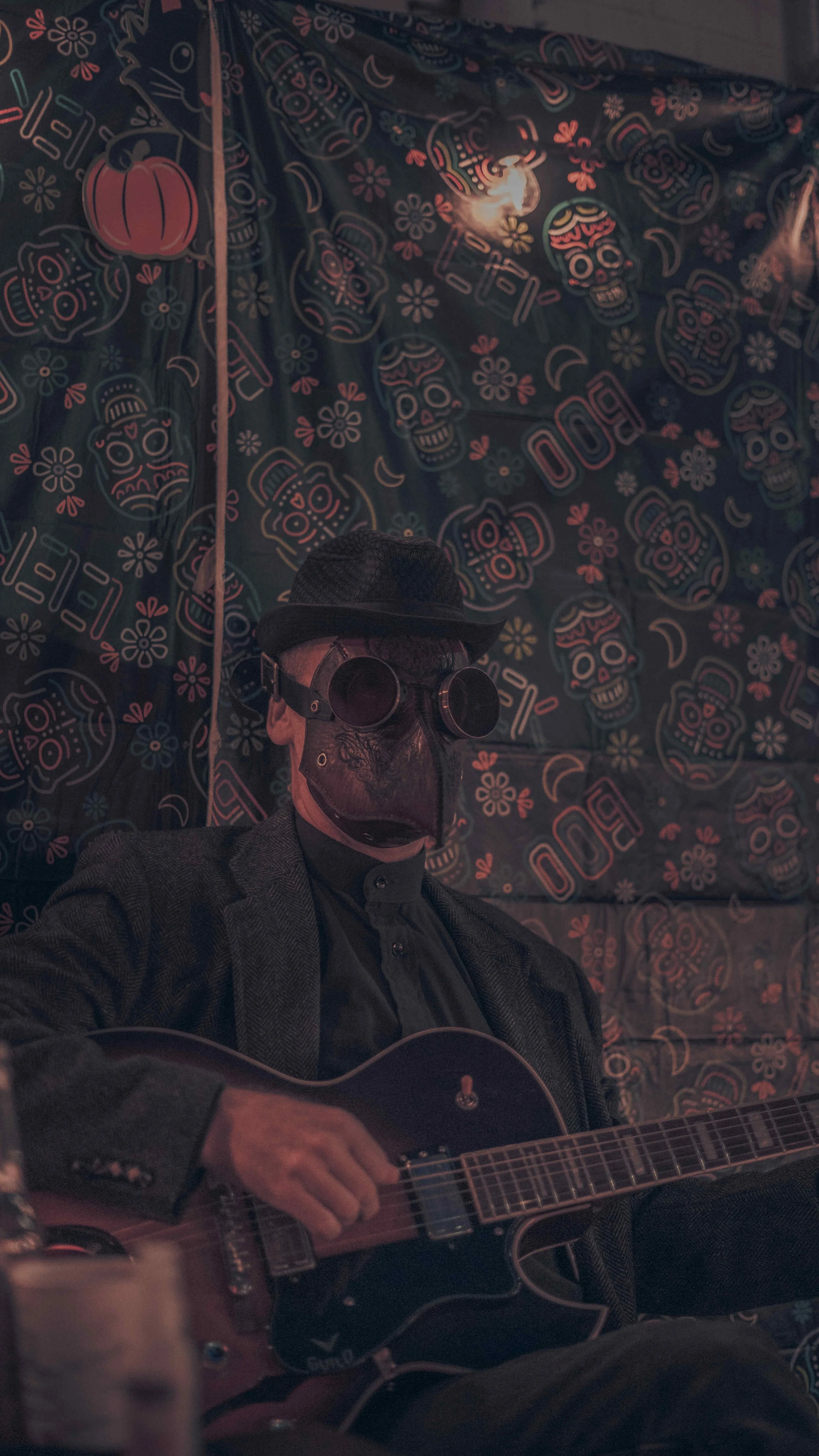 Person wearing a skull mask, sunglasses, and a black hat, playing an acoustic guitar in front of a wall decorated with Día de los Muertos skull and flower patterns.
