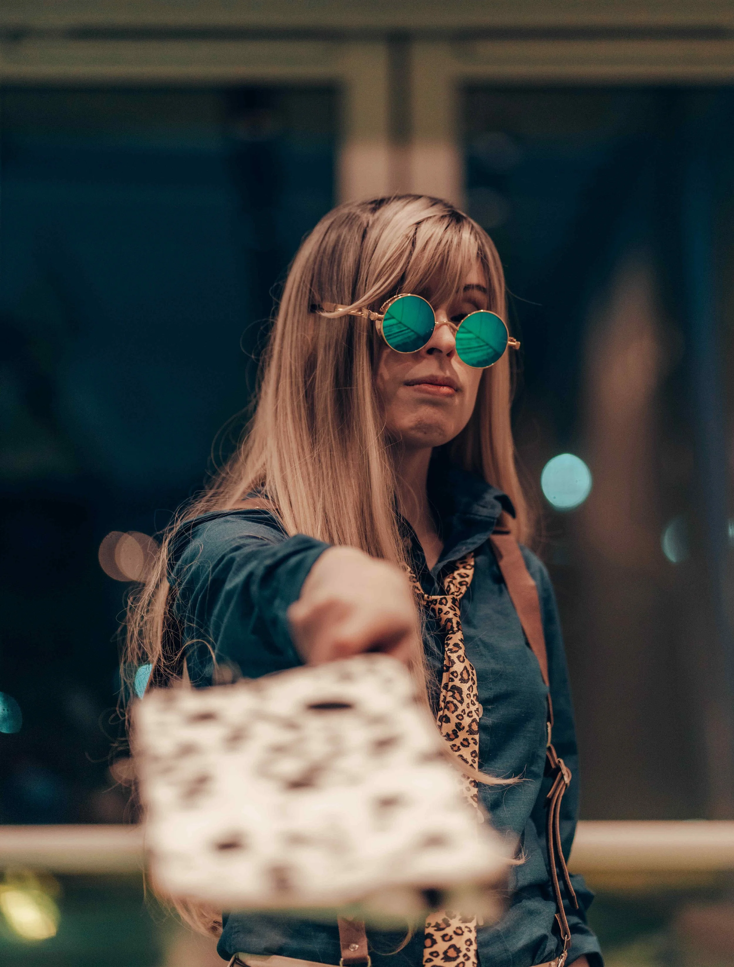 A woman with long blonde hair wearing round reflective sunglasses, a denim jacket, and a leopard print scarf, holding sushi in front of her.
