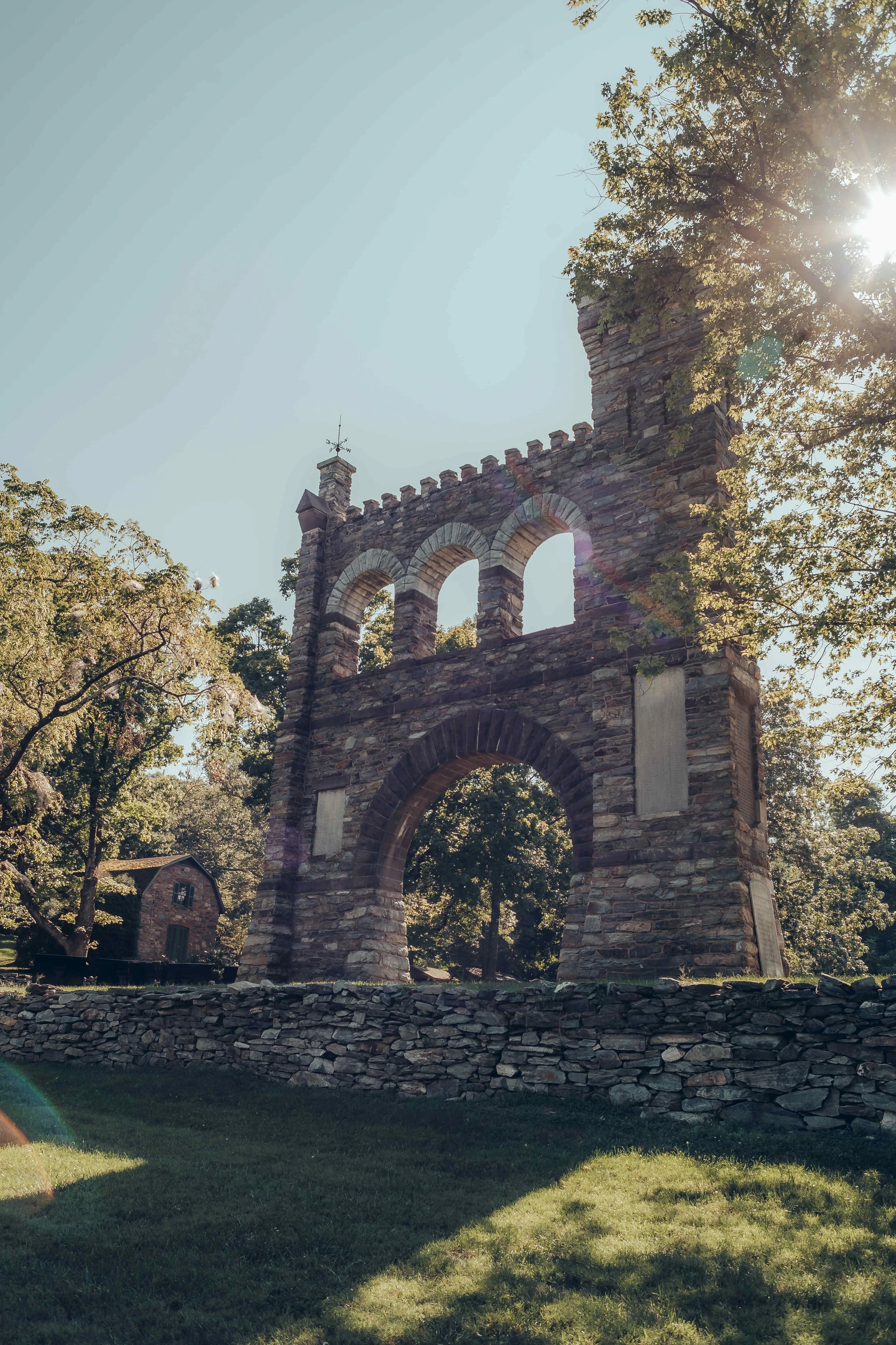 A historic stone archway with three arches, part of an old structure, surrounded by trees with green foliage, sunlight shining through the branches, and a clear sky in the background.