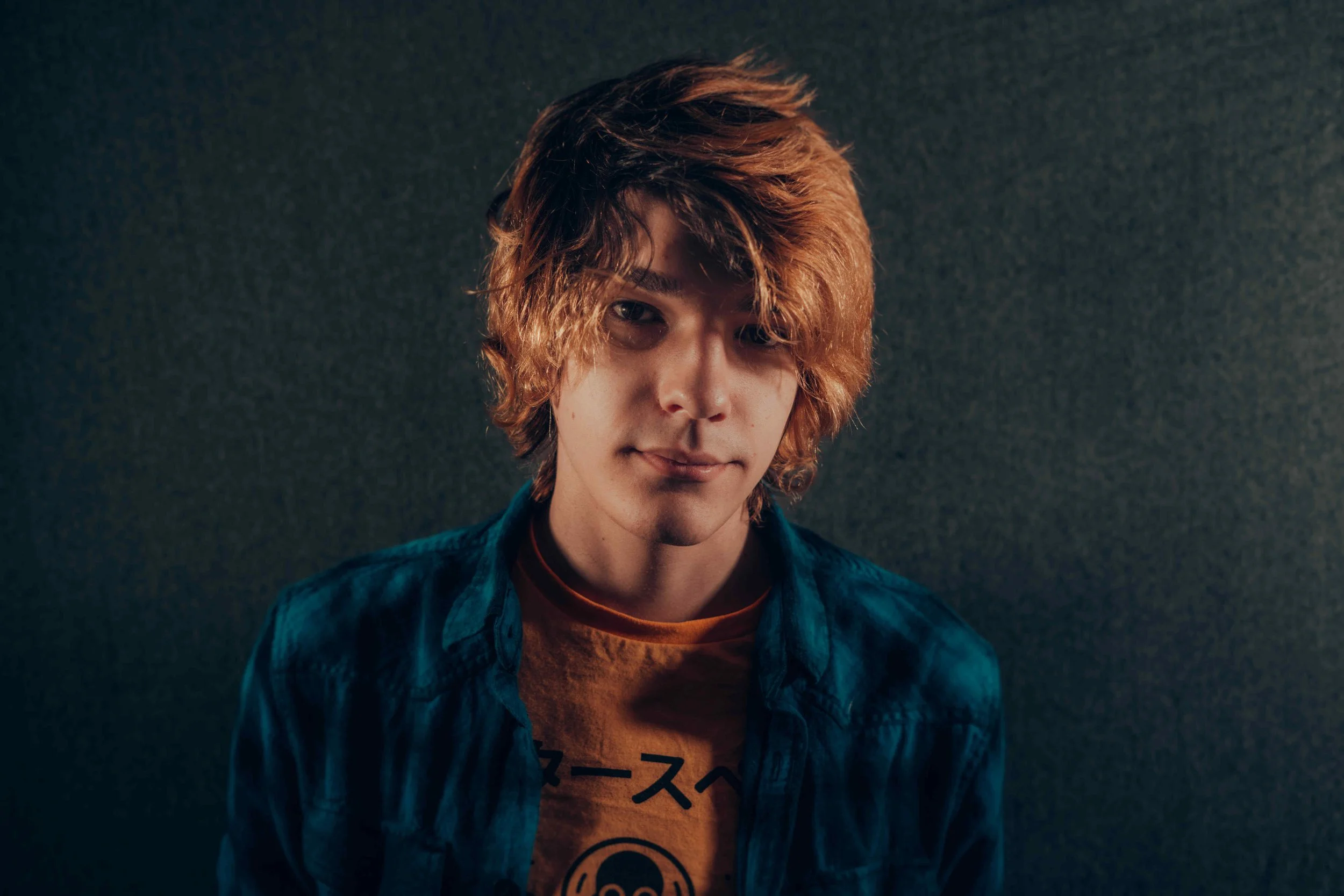 Young man with wavy, reddish-brown hair wearing a denim jacket over a graphic t-shirt, standing against a dark, textured background.