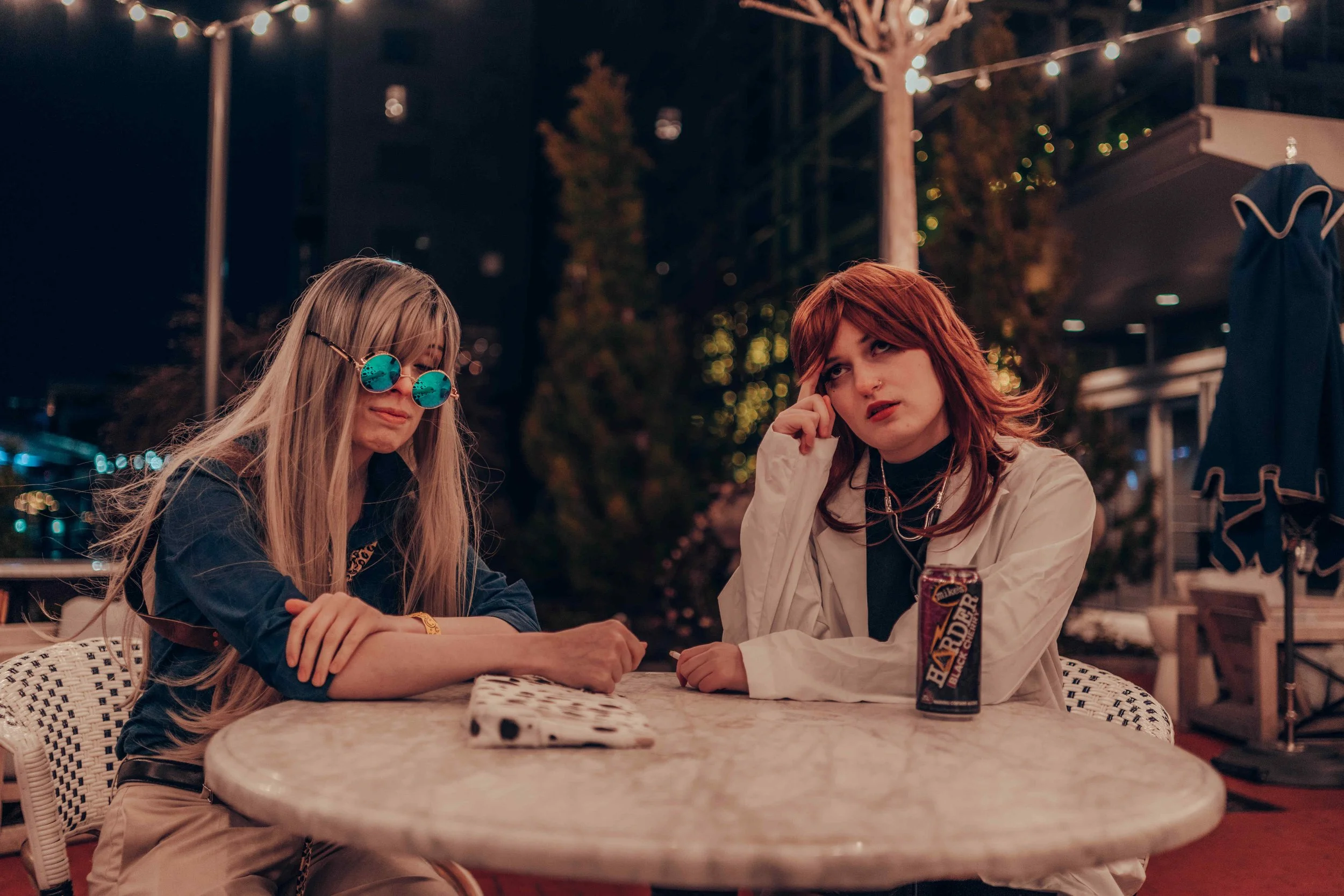 Two young women sitting at a table outdoors at night. One has long blonde hair, wearing reflective sunglasses, and the other has short red hair, wearing a beige jacket. They appear to be deep in conversation. There is a can of beer and a person in th