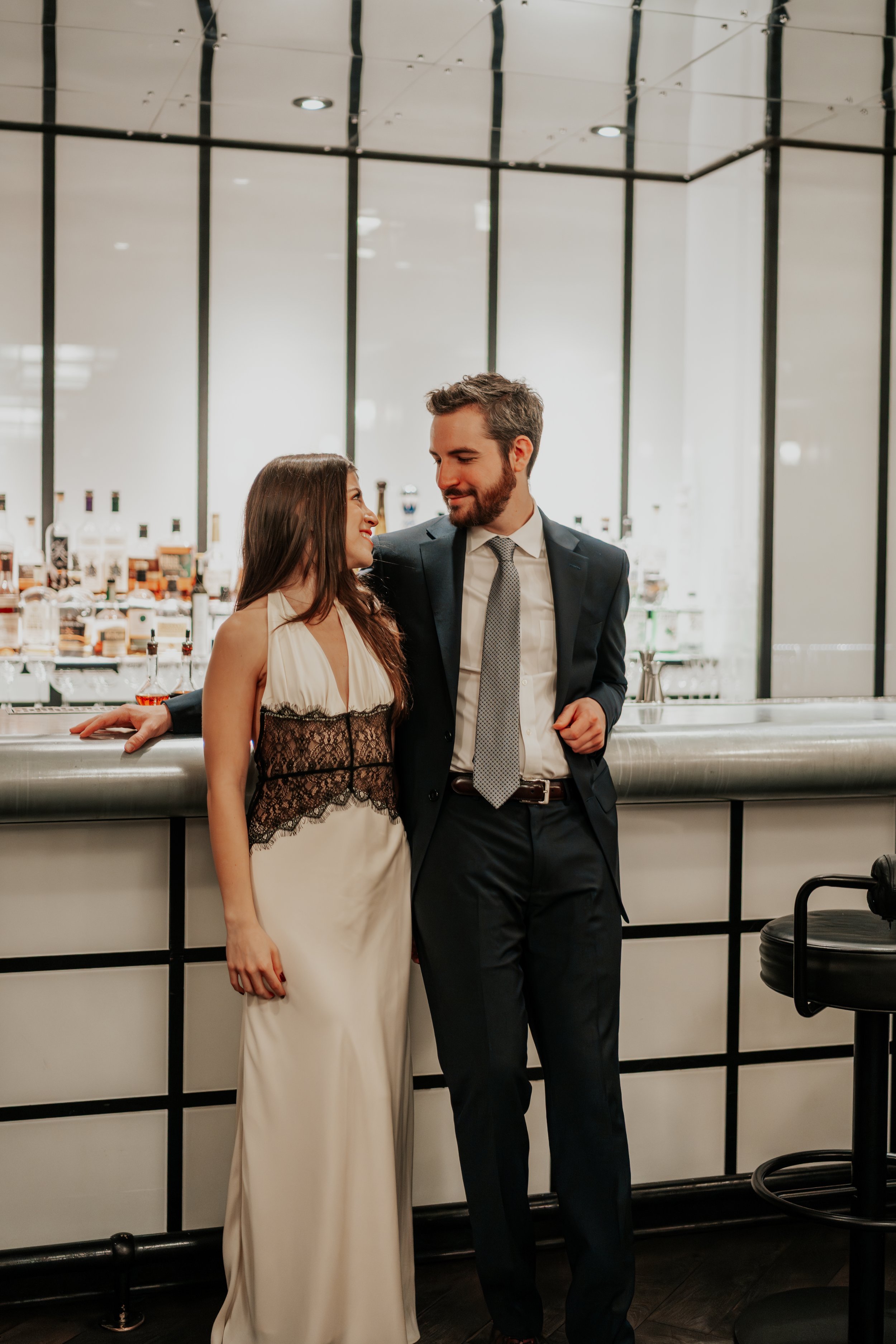 A man and woman in formal attire standing at a bar, gazing at each other with smiles in a modern, upscale setting.