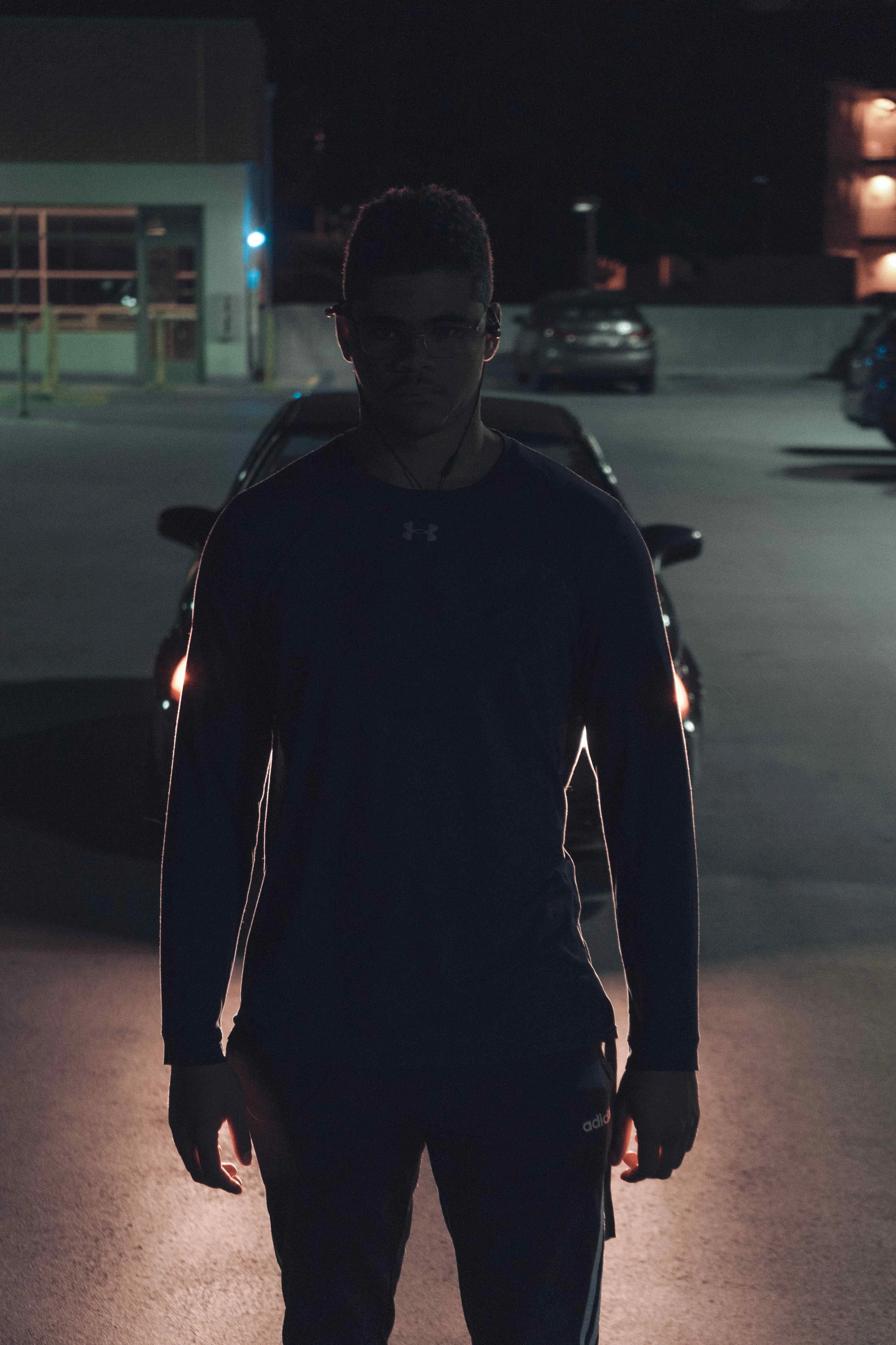 A person stands in a dark parking lot at night, illuminated from behind by streetlights, wearing glasses, a dark Under Armour shirt, and workout pants.