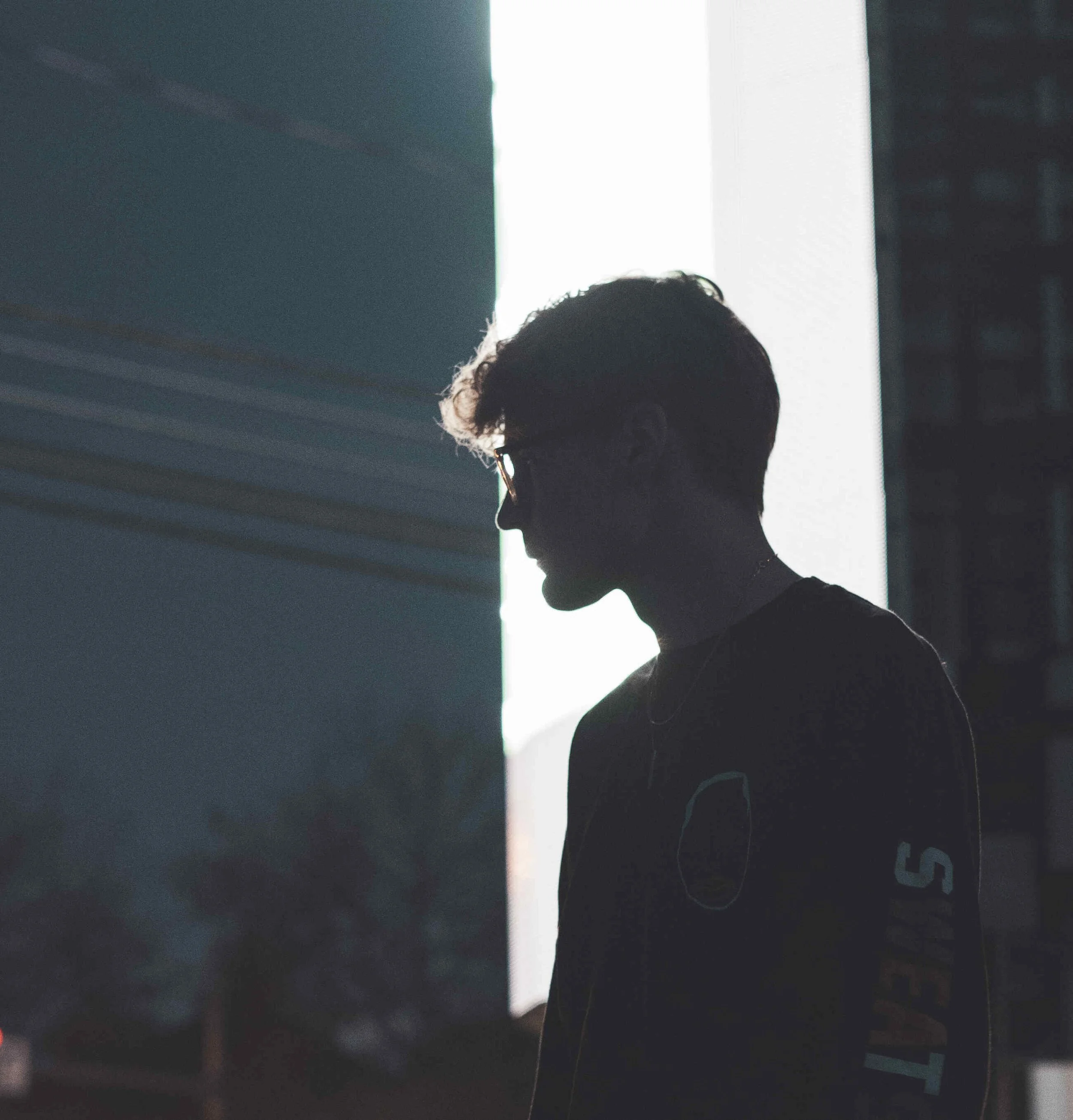 Silhouette of a person with glasses and curly hair, standing near a brightly lit window in a dark room.