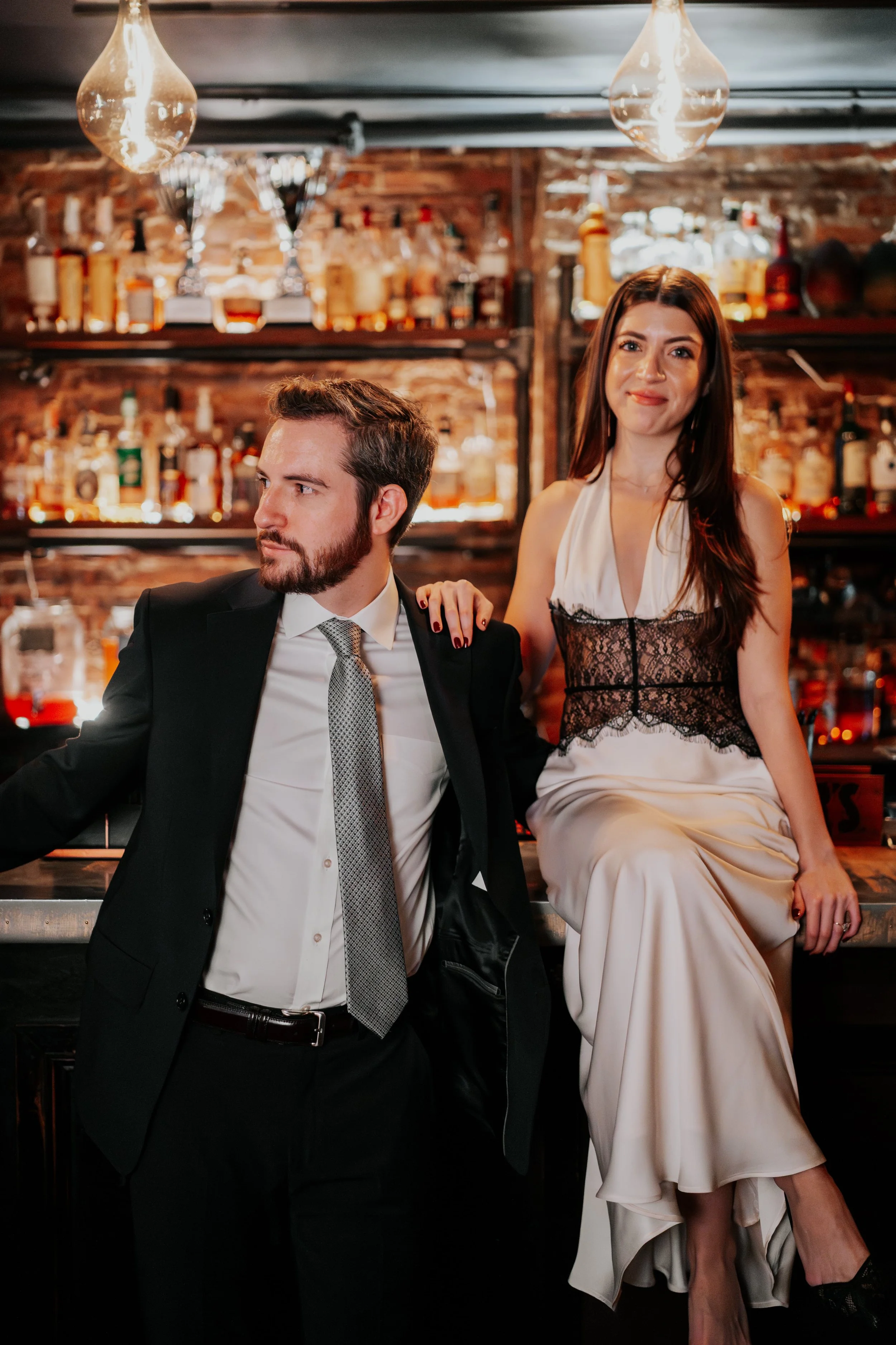 A man in a black suit with a gray tie and a woman in a white dress with black lace sitting at a bar with a brick wall and liquor bottles in the background.
