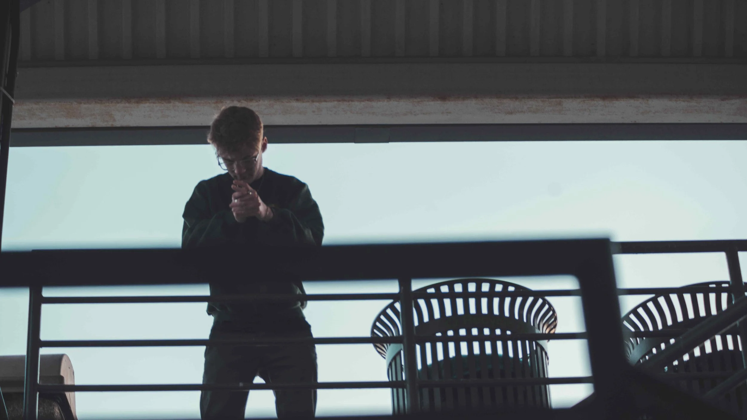A person standing on a balcony, looking down at a phone or device, with chairs and a railing in the foreground and a cloudy sky in the background.